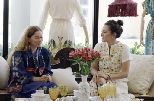 Two women sitting at a table with yellow drinks, engaged in conversation, in a bright room with floral decor and a dress on a mannequin in the background.