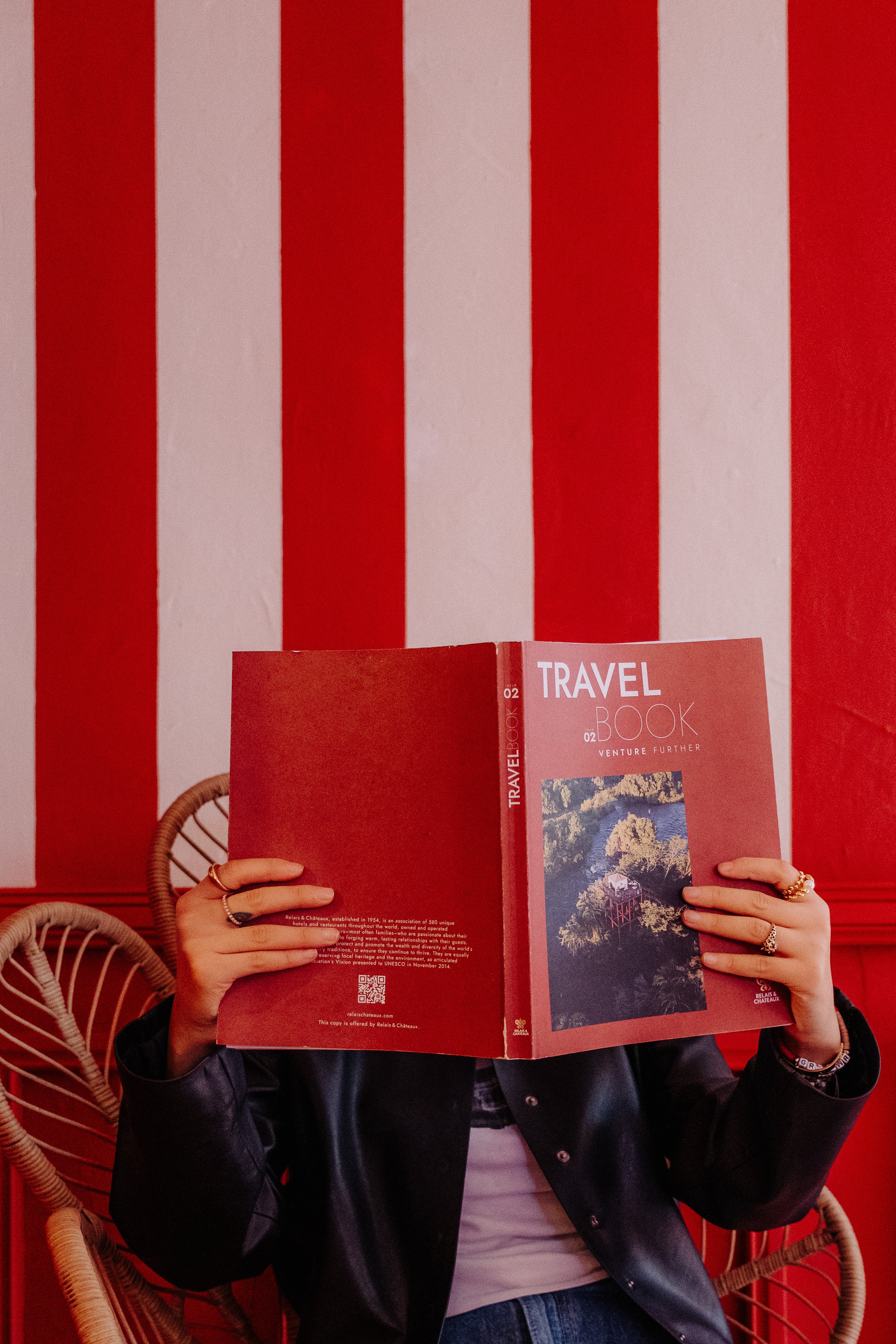 Person sitting in front of a red and white striped wall, holding a travel book in front of their face. The book cover shows a mountainous landscape.