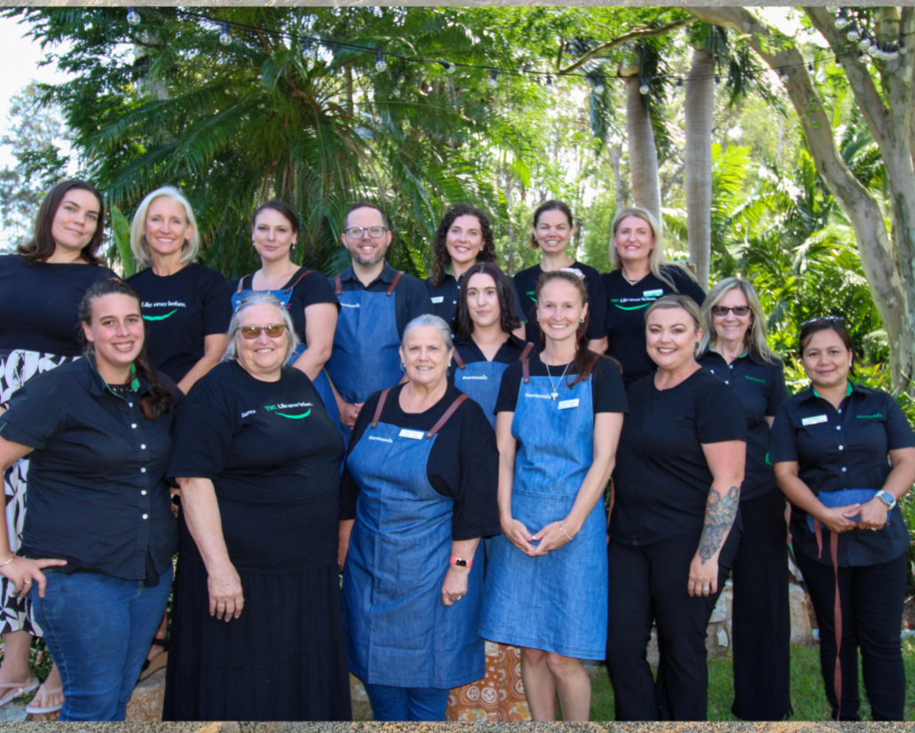 Group of fifteen people, mostly women, standing outdoors in a tropical garden with lush green trees and plants, some wearing black shirts and others in blue aprons, smiling for a photo.