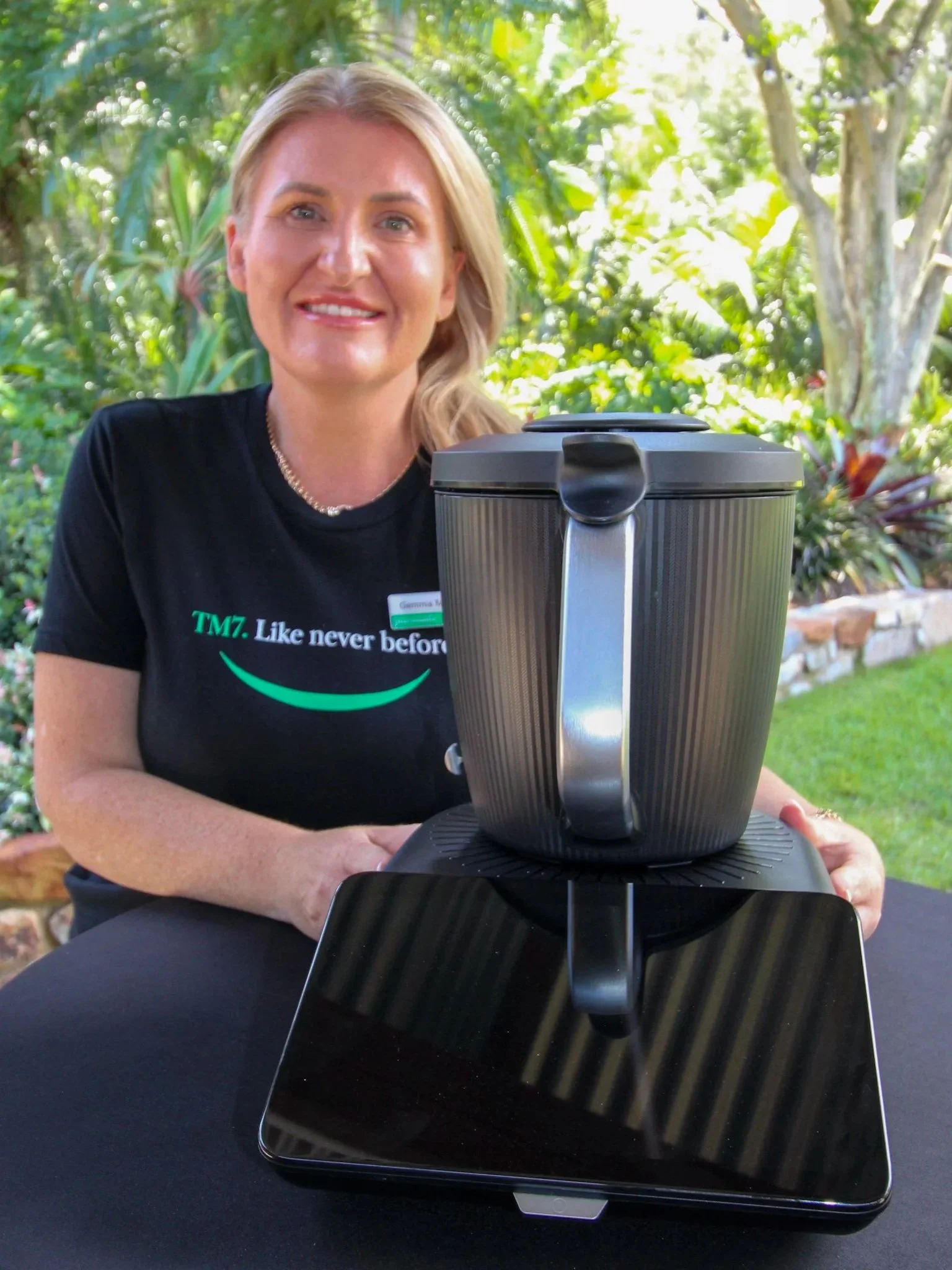 A smiling woman with blonde hair outdoors, sitting at a table with a black cloth, a black tablet, and a coffee pot.
