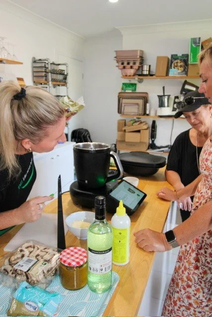 Three women in a kitchen looking at a tablet on the counter with various kitchen items and supplies around them.