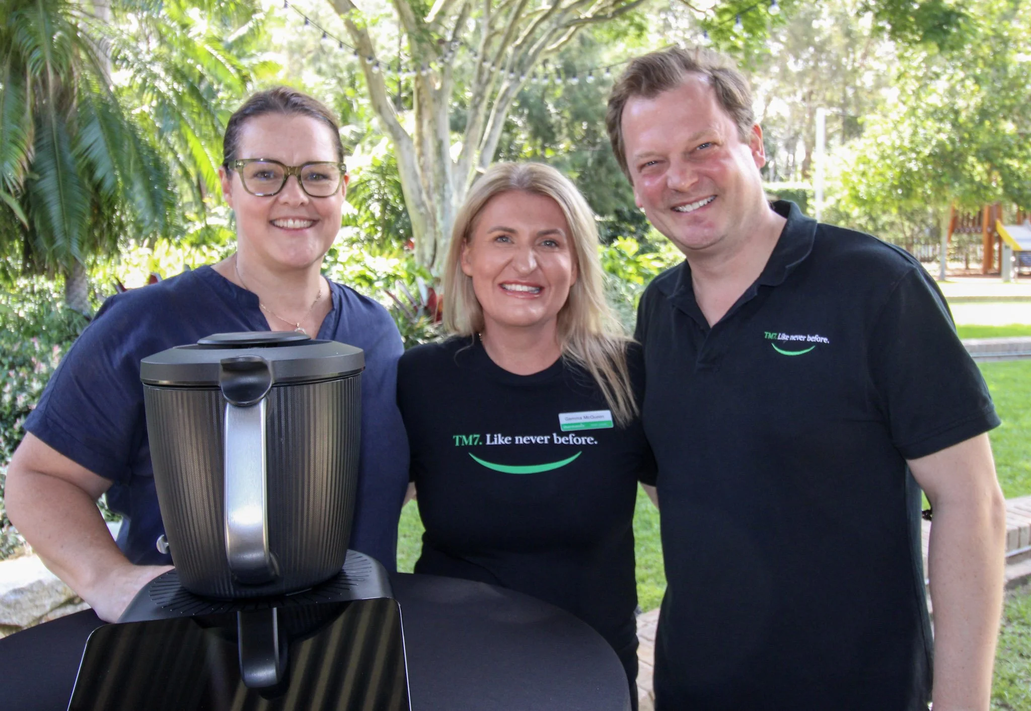 Three smiling people standing together outdoors with trees and grass in the background. One woman is wearing glasses and a navy shirt, standing next to a black trash can or large container. The middle woman has blonde hair and is wearing a black T-shirt with the words "TM7. Like never before." The man on the right has light brown hair and is wearing a black polo shirt with similar branding.