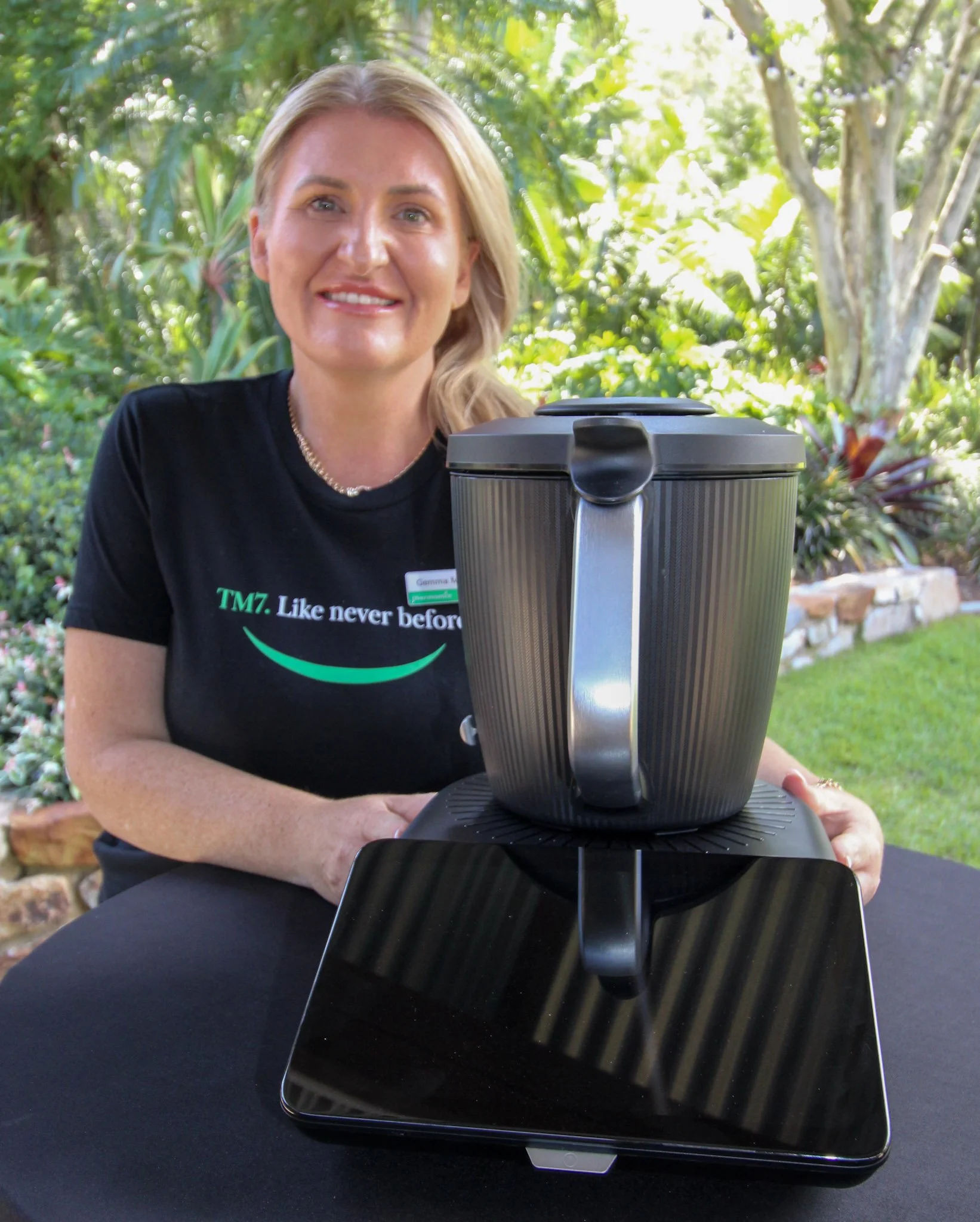 A woman sitting outdoors at a table with a large black plastic container and a black tablet in front of her, surrounded by greenery and plants.