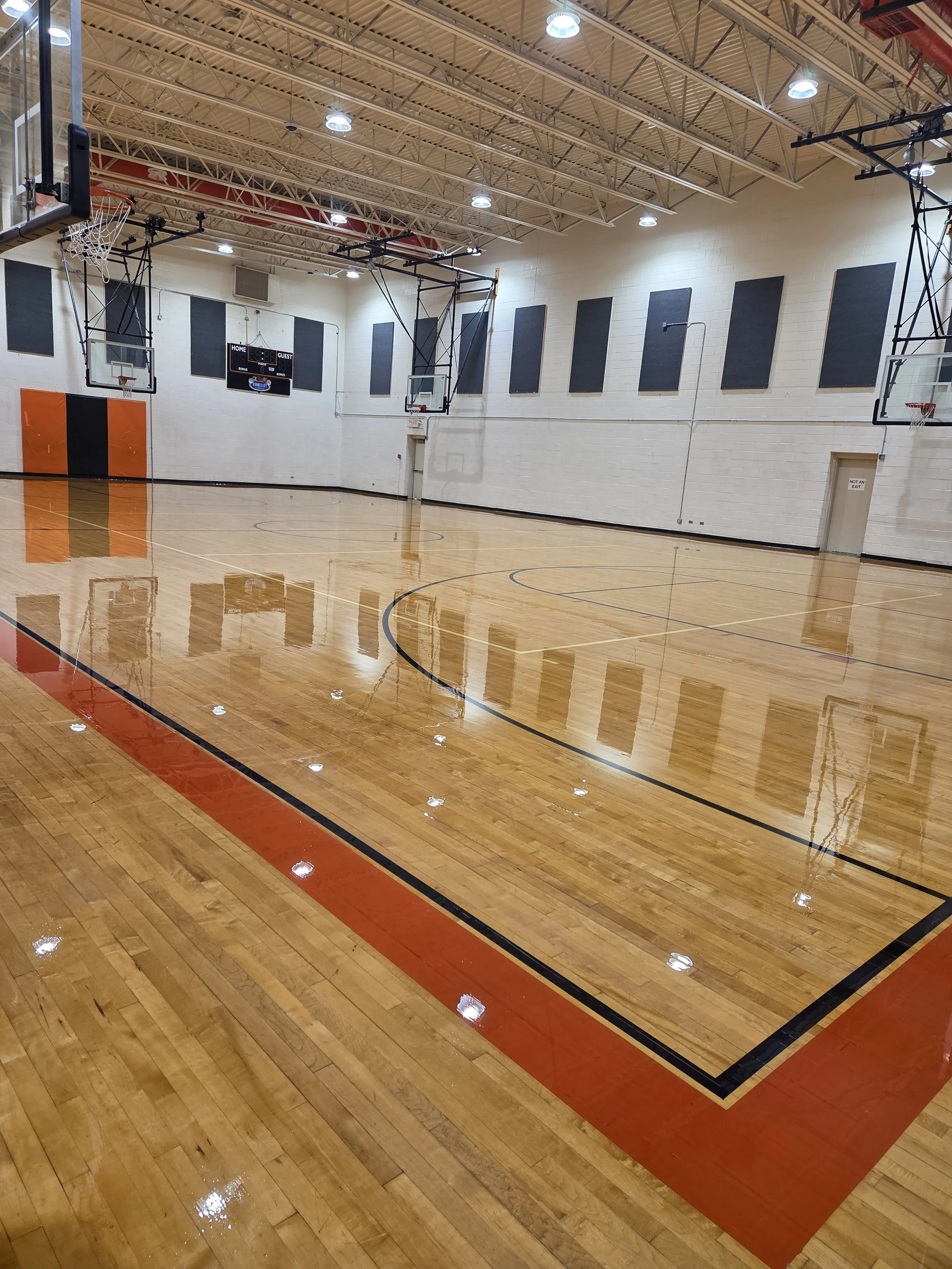 An indoor basketball gymnasium with polished wooden flooring, multiple basketball hoops, a scoreboard, and black wall-mounted acoustic panels.