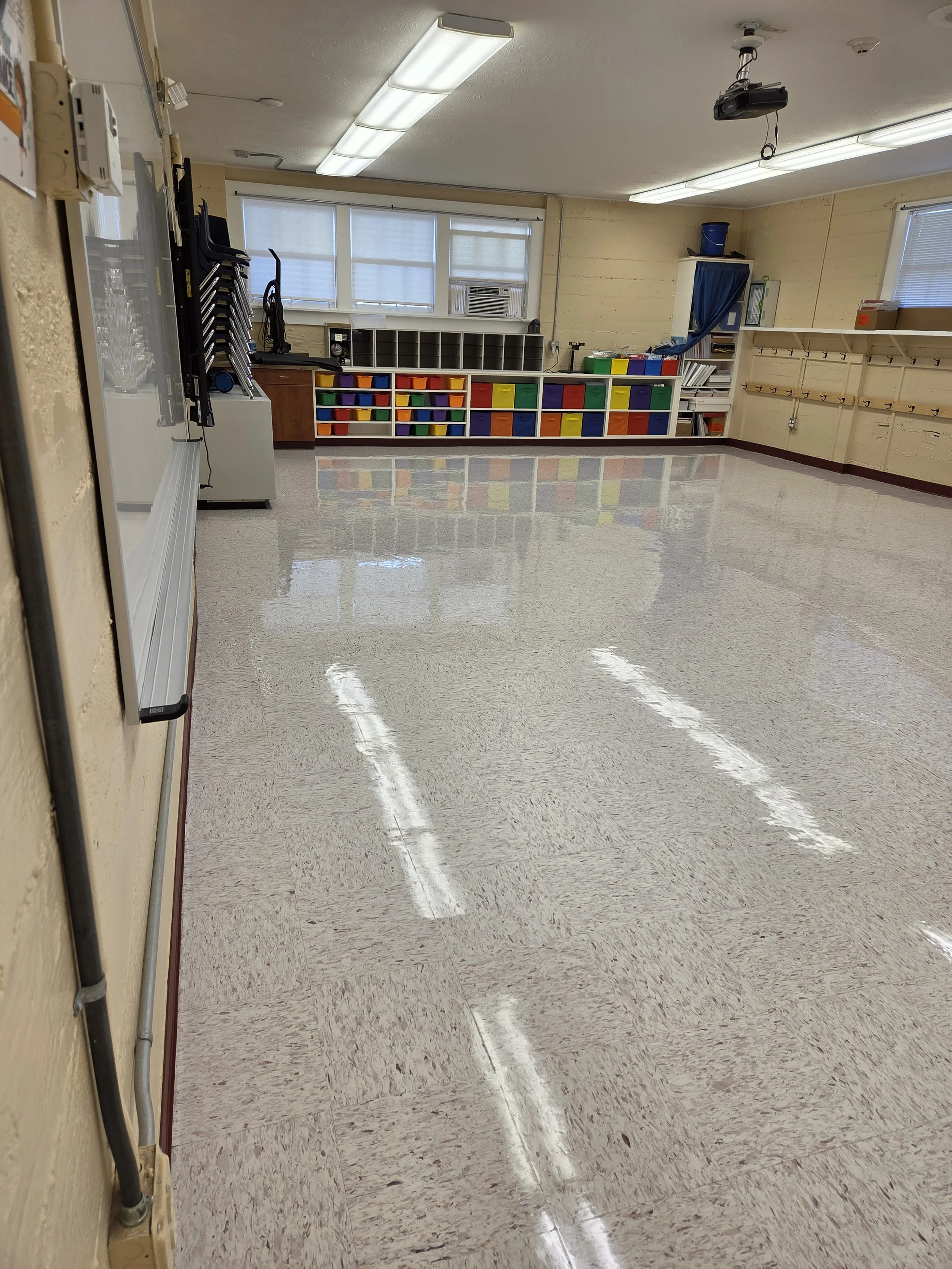 Empty classroom with a shiny tile floor, a whiteboard on the left, colorful storage bins on the back shelf, and fluorescent lights on the ceiling.