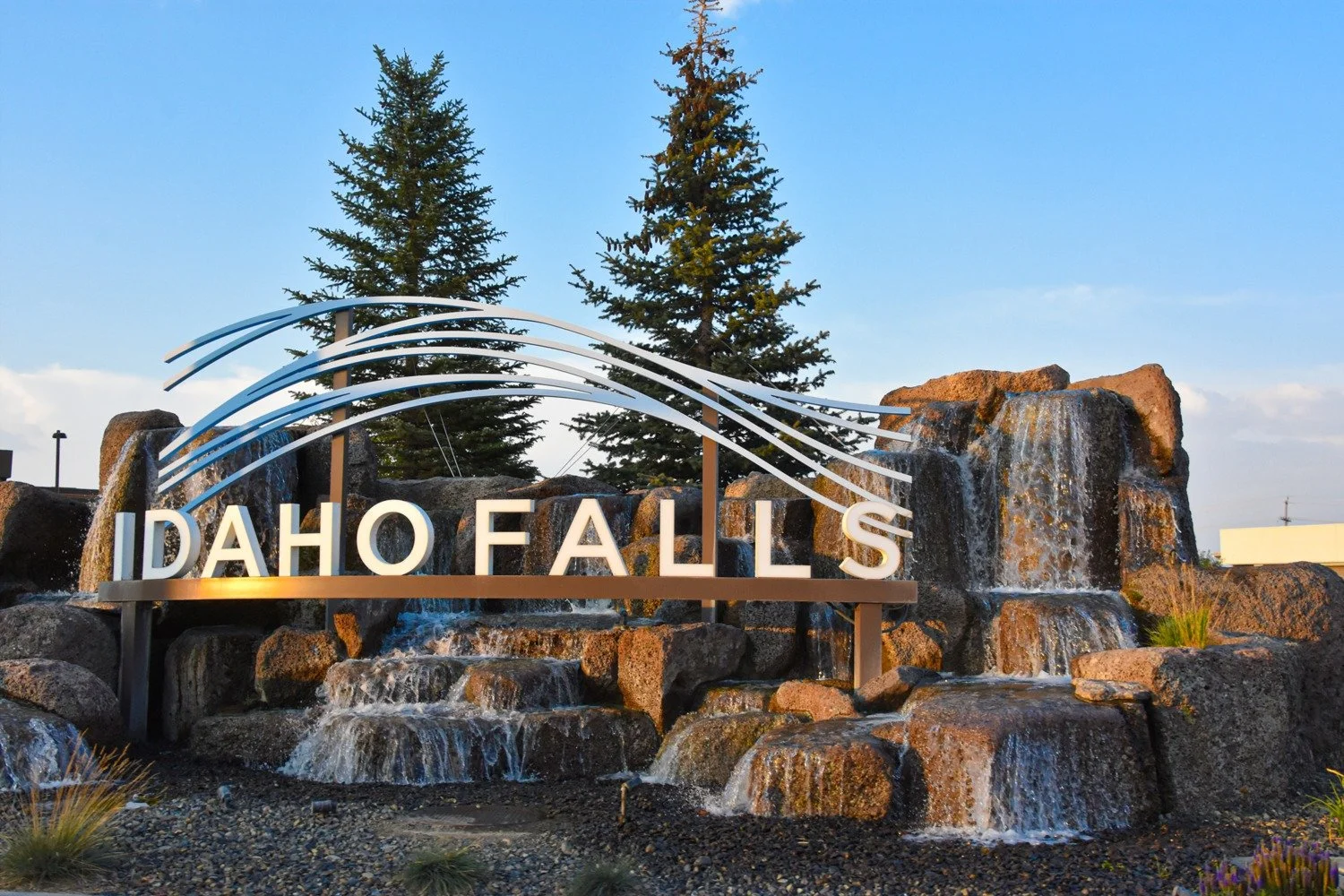 Sign at Idaho Falls with water feature and waterfalls, trees, and a clear sky in the background.