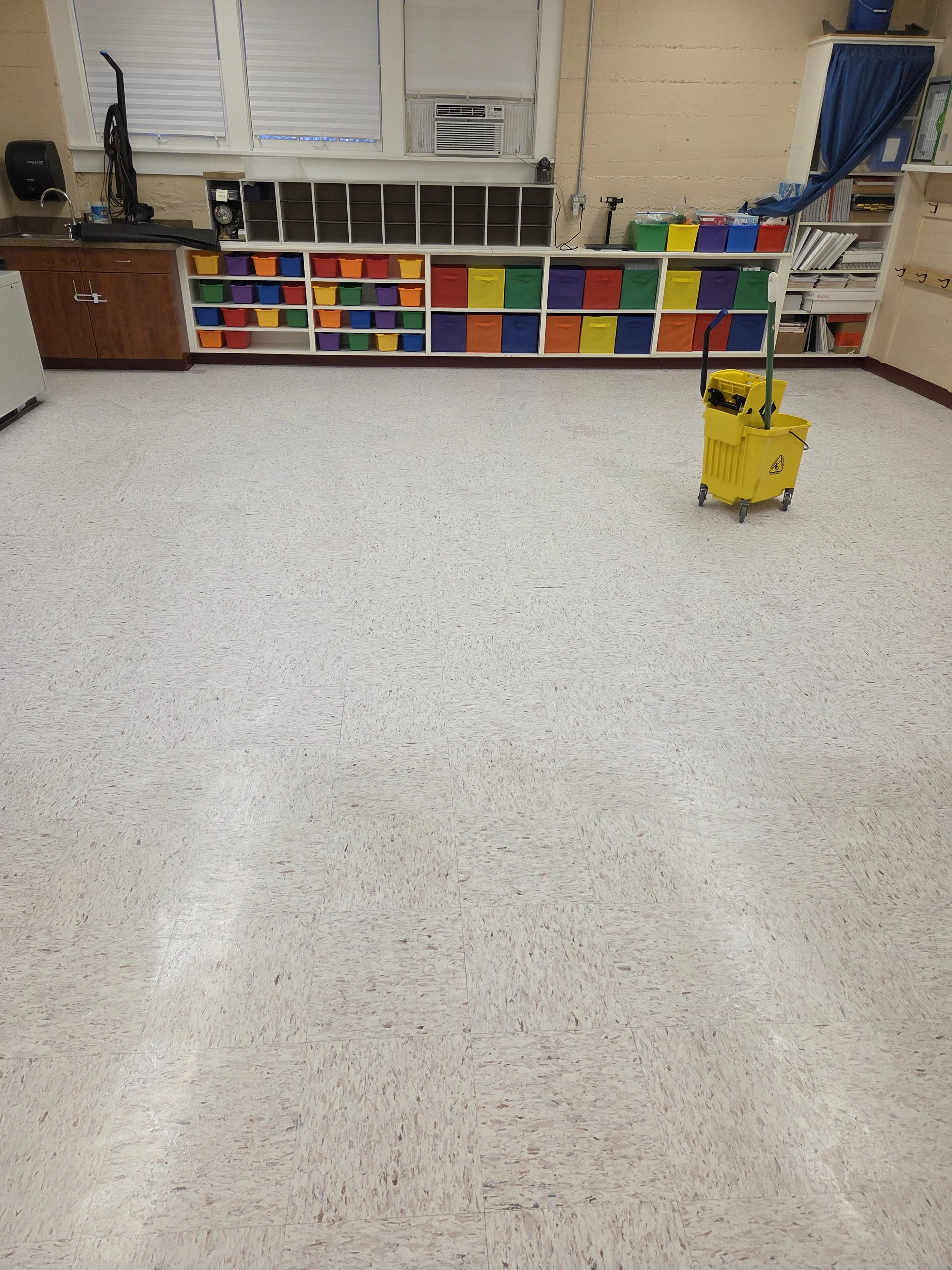 Empty classroom with tiled floor, colorful cubbies, a mop and bucket, and shelves with books and supplies.