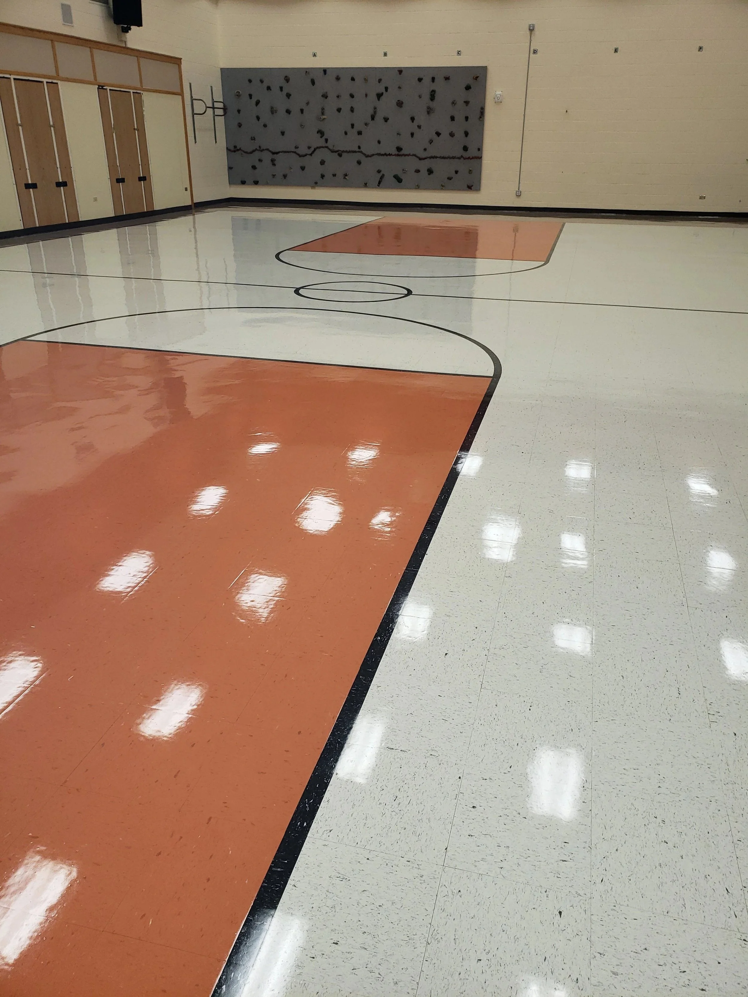 Empty gymnasium with a climbing wall on the back wall and a basketball court with orange and white floor surfaces.