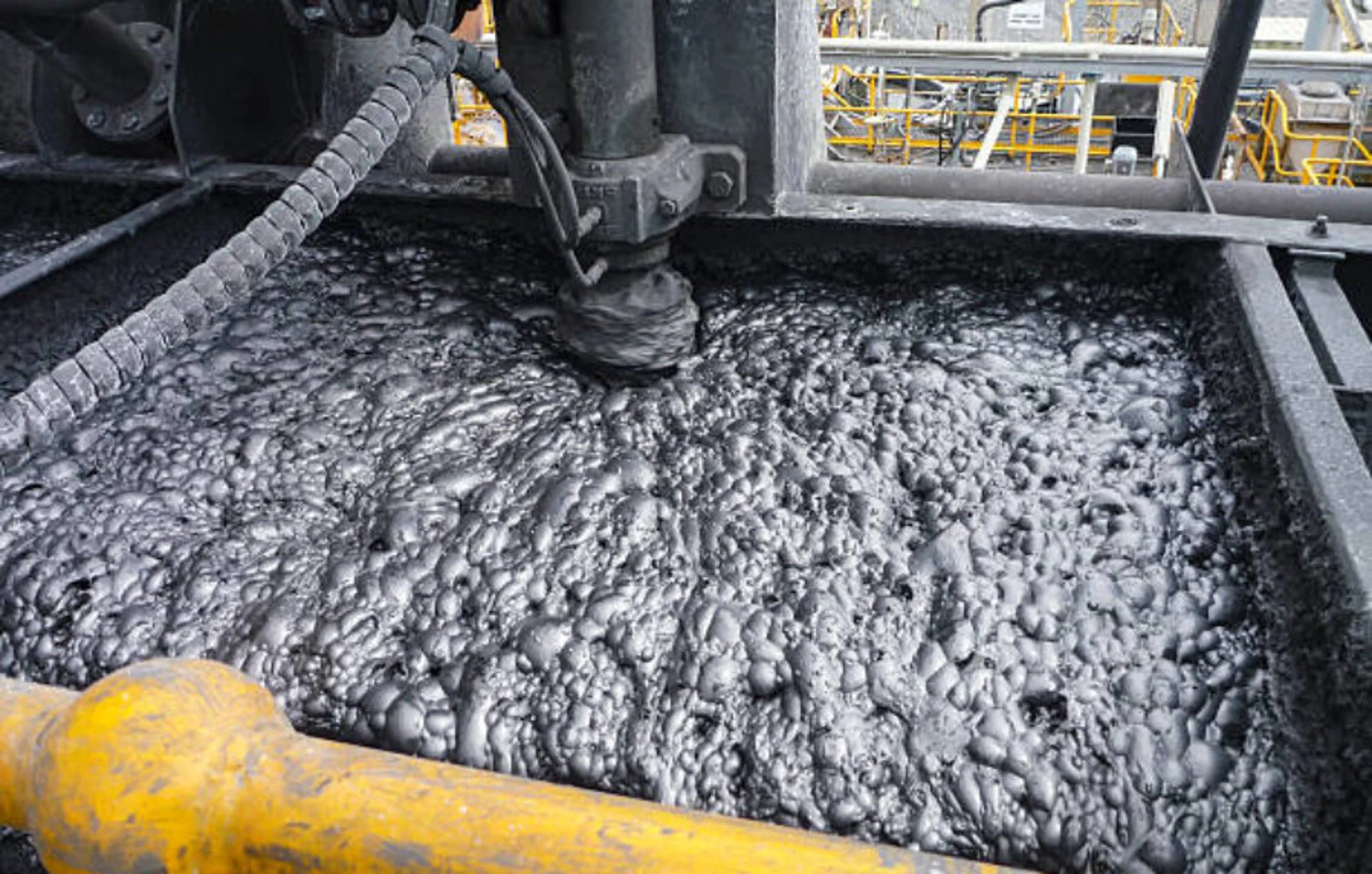 Industrial process with black foamy liquid in a large container, surrounded by yellow and black safety barriers, with equipment and industrial structures in the background.