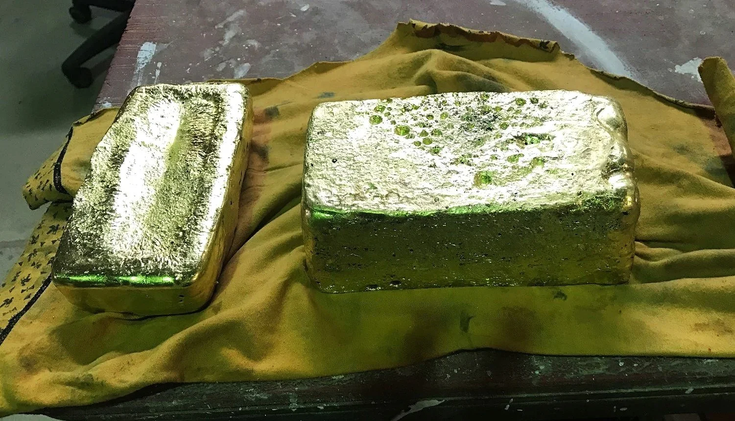Two gold-colored metal ingots resting on a yellow cloth on a dark surface.