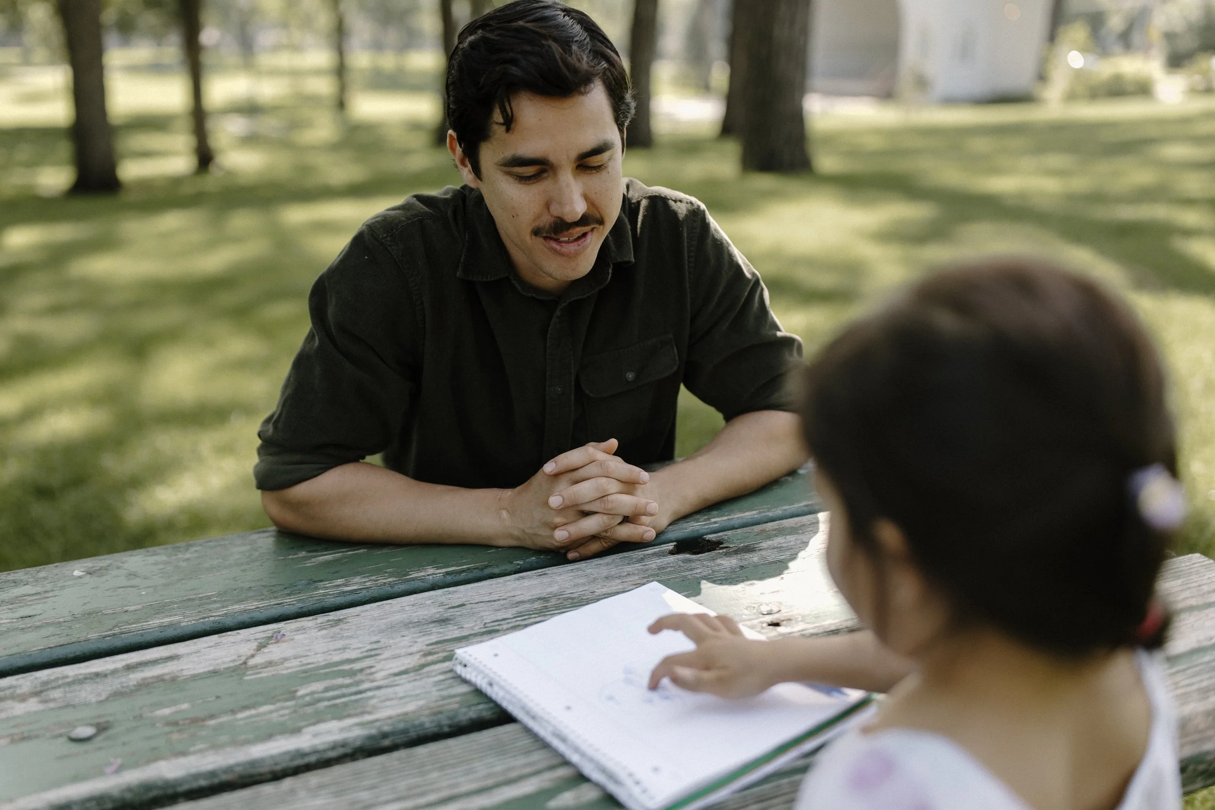 A man with dark hair and a mustache, wearing a black shirt, is sitting at a picnic table in a park, talking to a child. The child is a girl with dark hair and a floral dress. They are outdoors with trees and grass in the background.