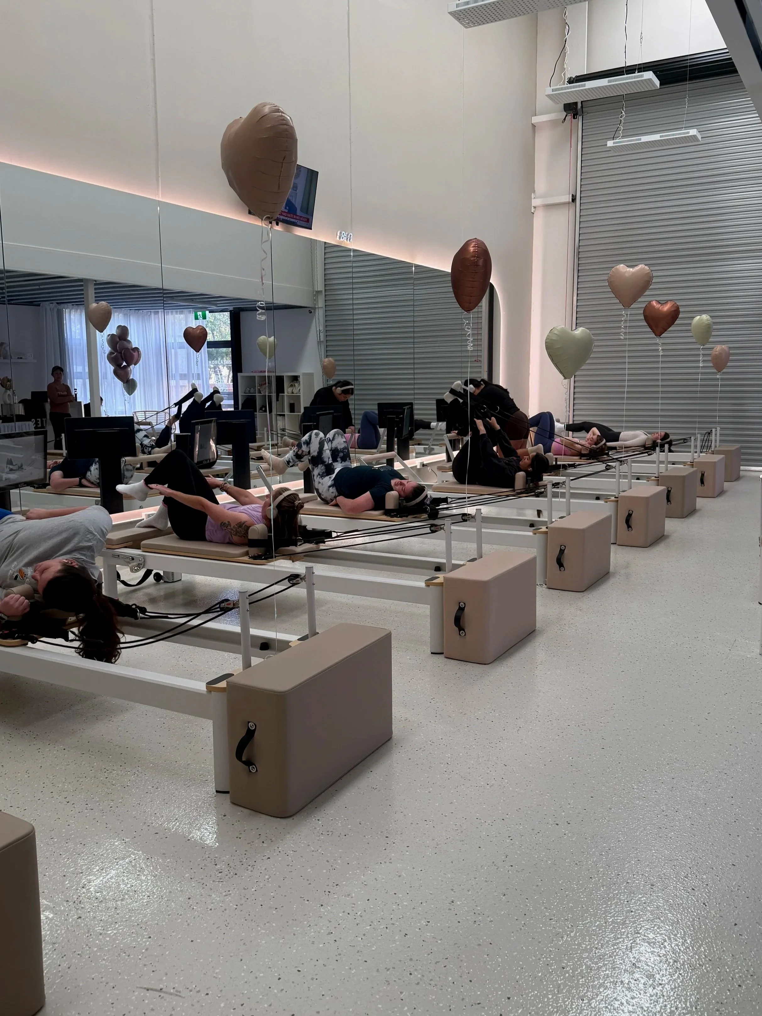 A fitness or physical therapy clinic with multiple women exercising on specialized Pilates reformer machines in front of a mirror, balloons hanging from the ceiling, and a television on the wall.