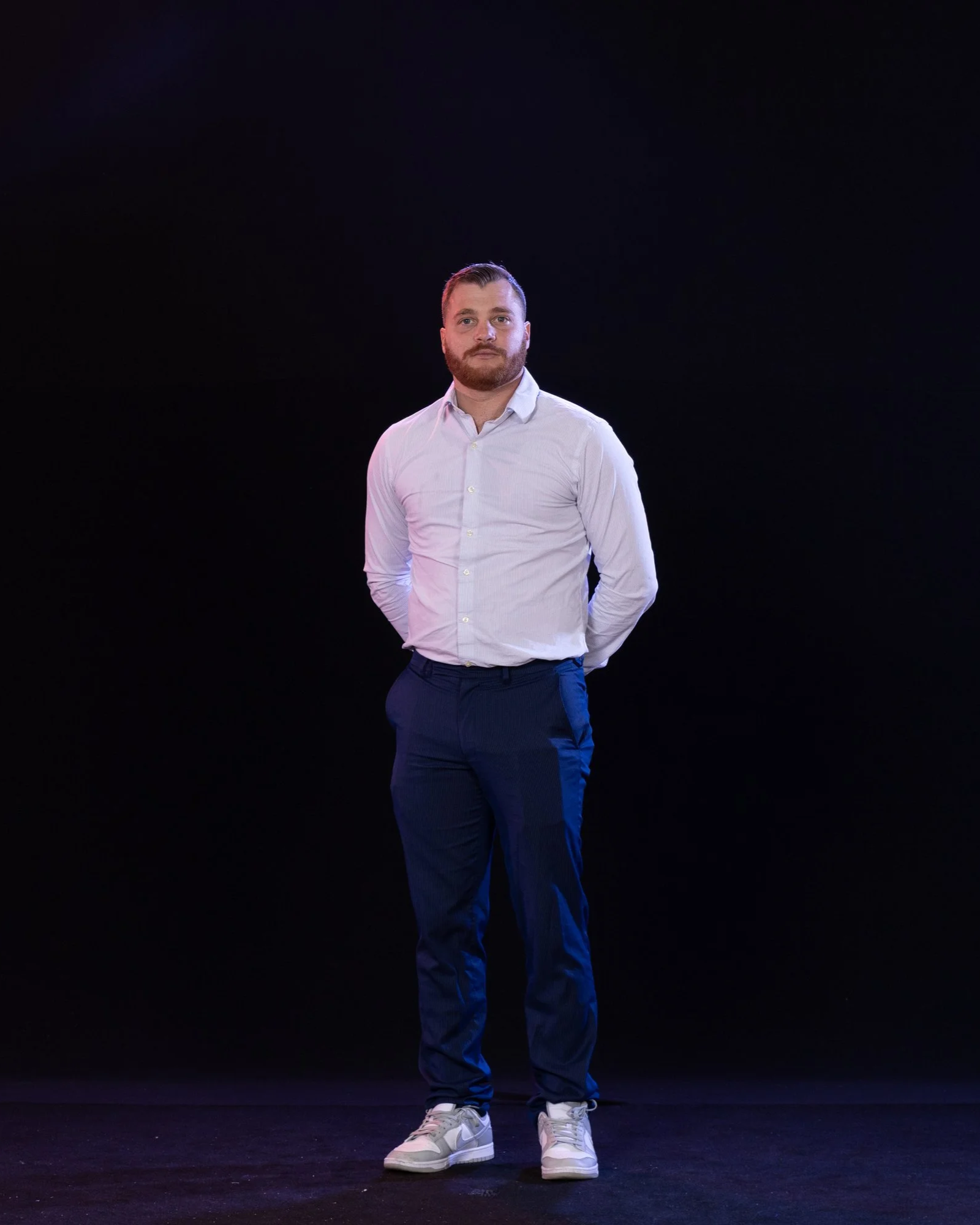Man with a beard in a white dress shirt and dark pants, standing on a dark stage with a black background, wearing white sneakers.