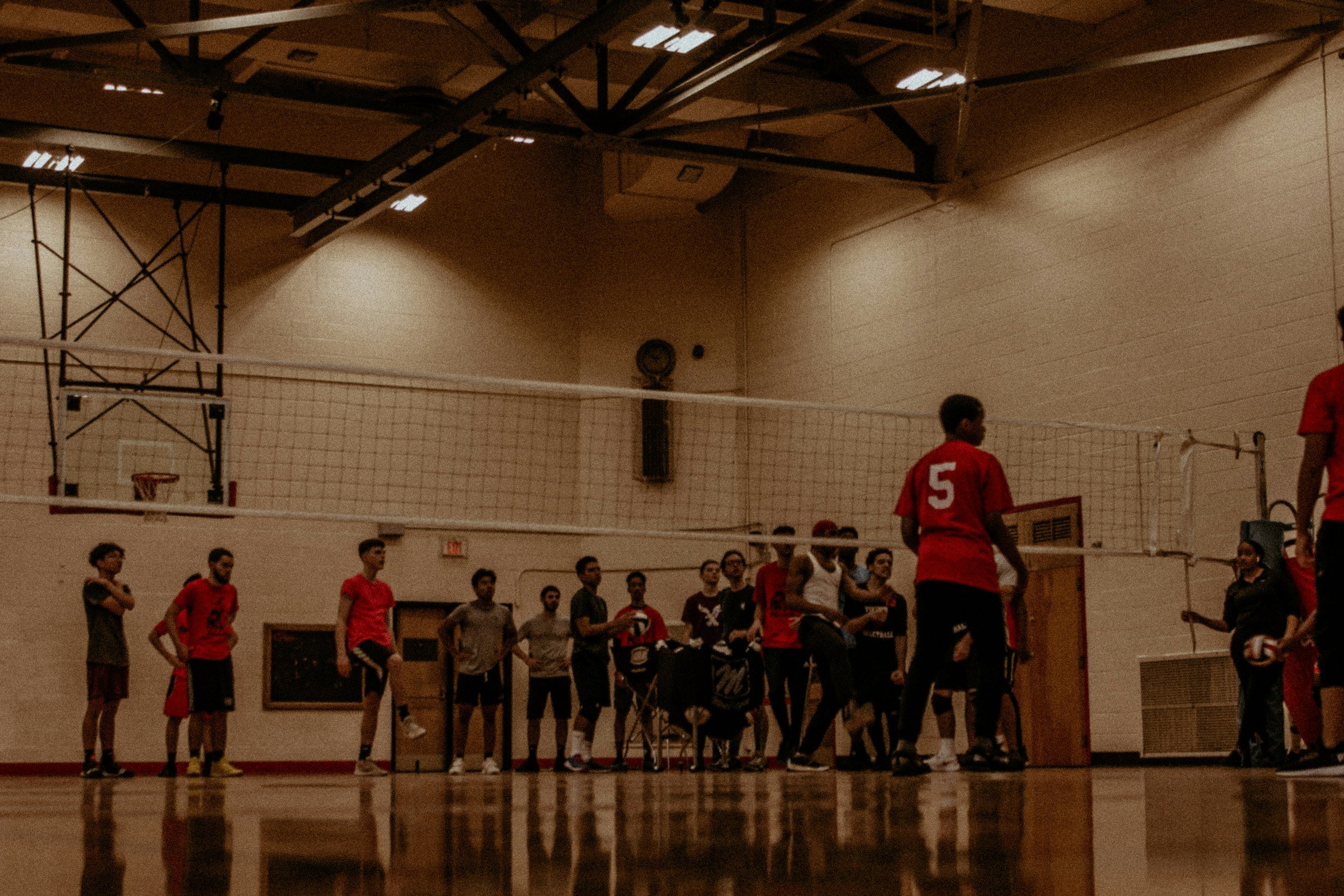 Indoor volleyball gym with a group of players standing near the net, with some wearing red jerseys, and others in gray or black clothes. The gym has a high ceiling with lighting fixtures and a basketball hoop on the side wall.