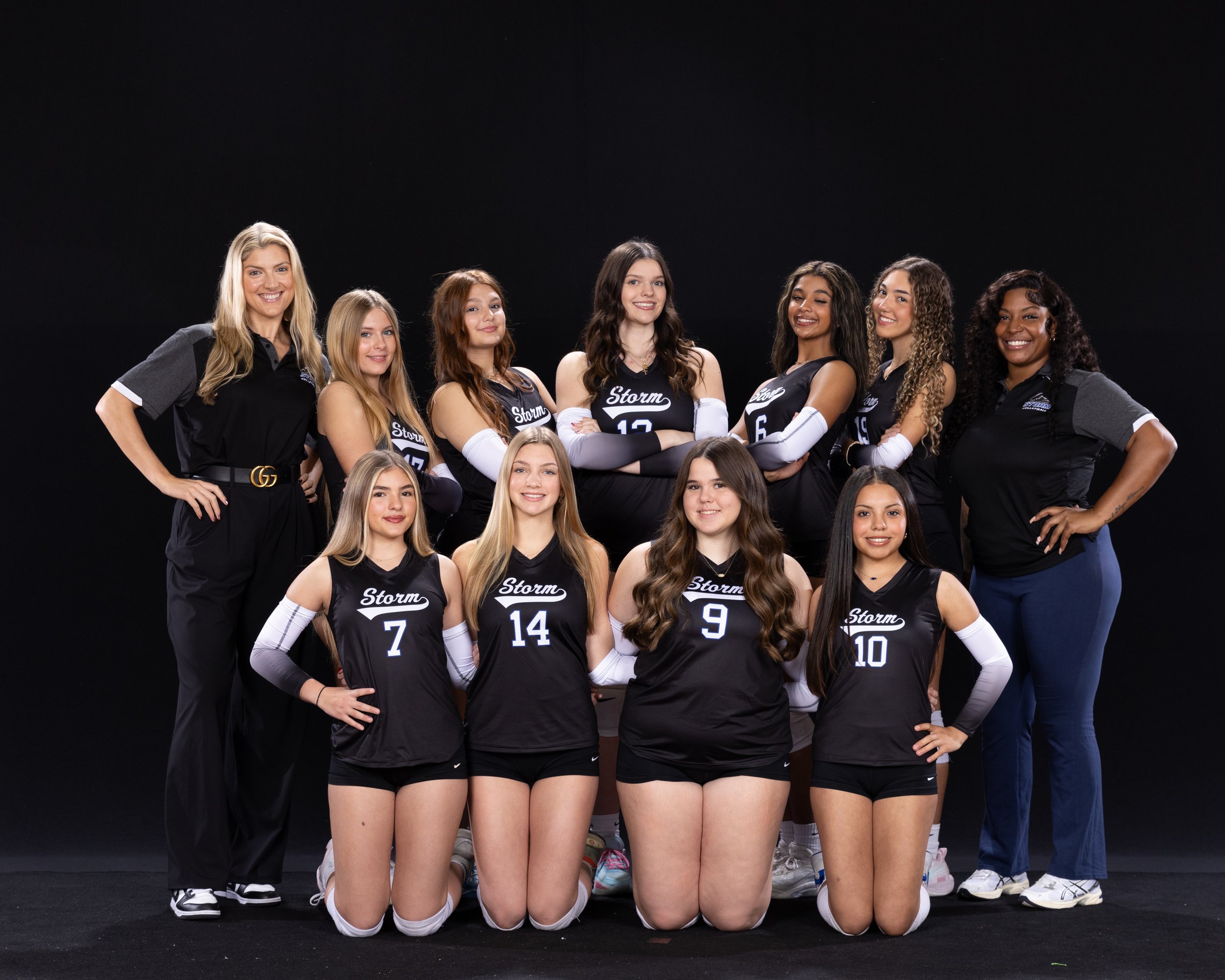 A group of volleyball players and coaches posing for a team photo against a black background. The players are wearing black and white uniforms with the word 'Storm' and their jersey numbers.