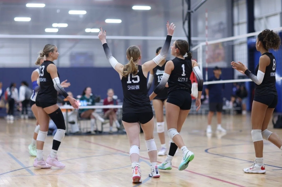Girls volleyball team celebrating on indoor court, some players with arms raised, others clapping, volleyball net in background, spectators at side.