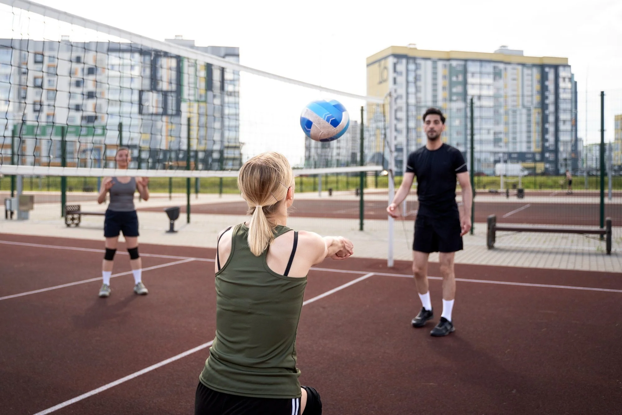 People playing volleyball on an outdoor court in a city with tall apartment buildings in the background.