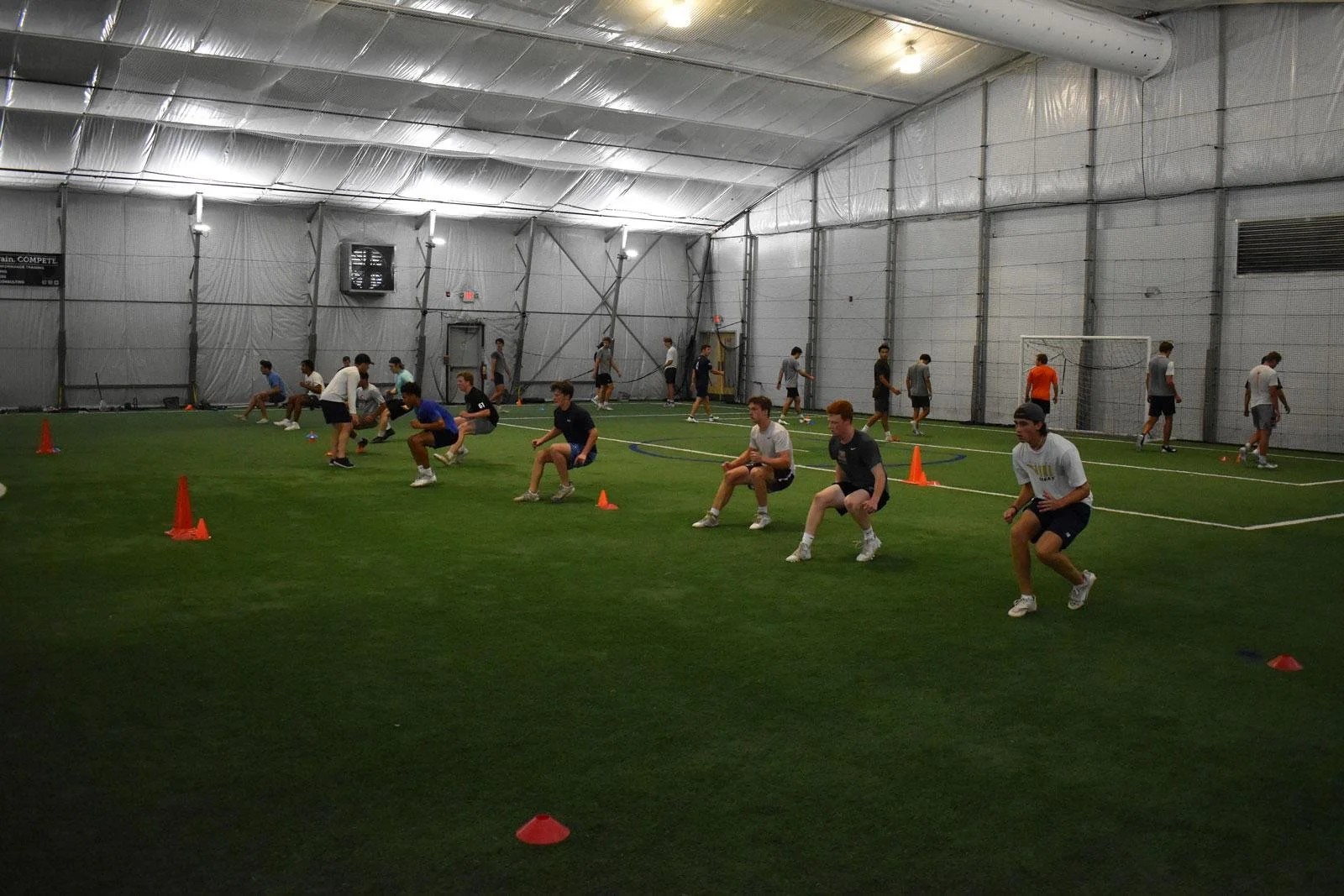 Young athletes working out inside an indoor sports facility with artificial turf, orange cones, and team training activity.