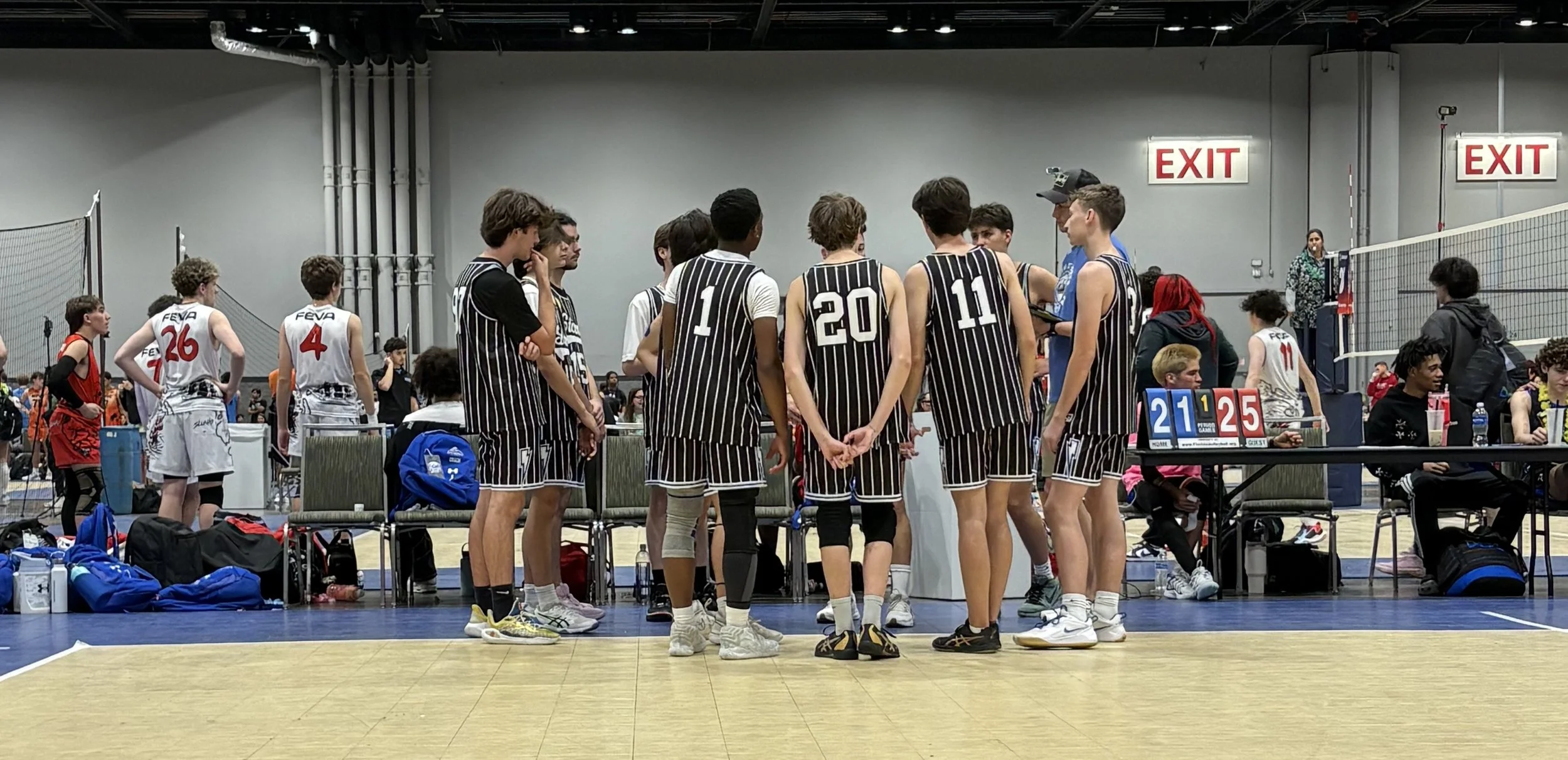 A group of young basketball players in black and white uniforms are gathered in the middle of an indoor basketball court, with some standing and some sitting nearby. In the background, there are other players, spectators, and scoreboards displaying a score of 21 to 25.