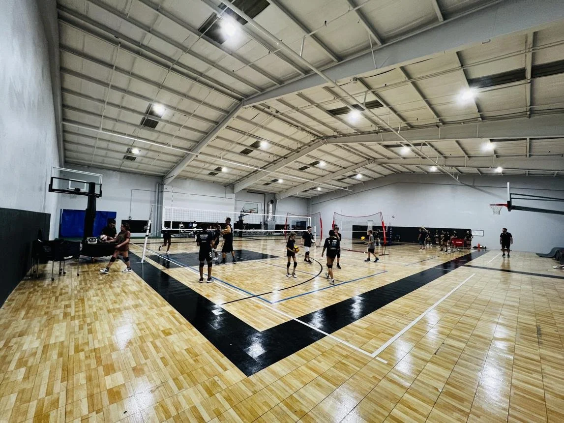 Indoor gym with volleyball and basketball activities, multiple players on court, sports equipment, and a high ceiling.