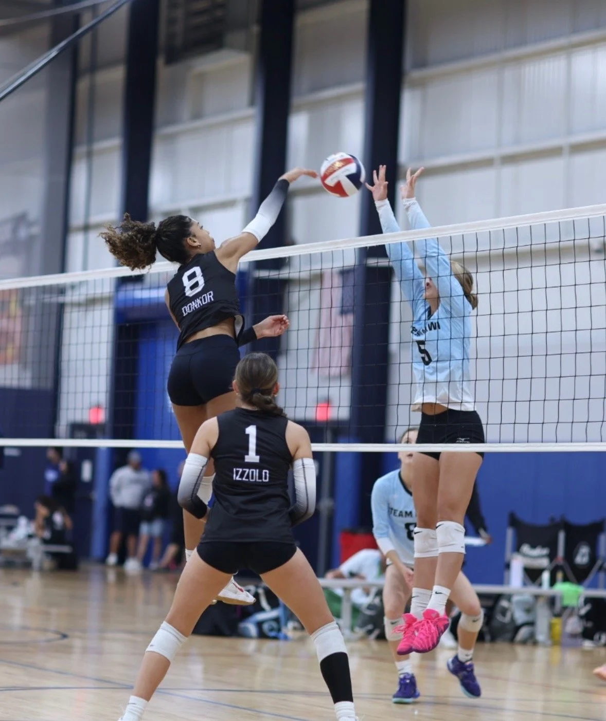 Two female volleyball players are jumping at the net; one in black jersey with the name 'DONKOR' and number 8, is attacking the ball, and the other in a light blue jersey with the number 5 is blocking. A third player in black with number 1, labeled 'IZZOLO,' is watching.
