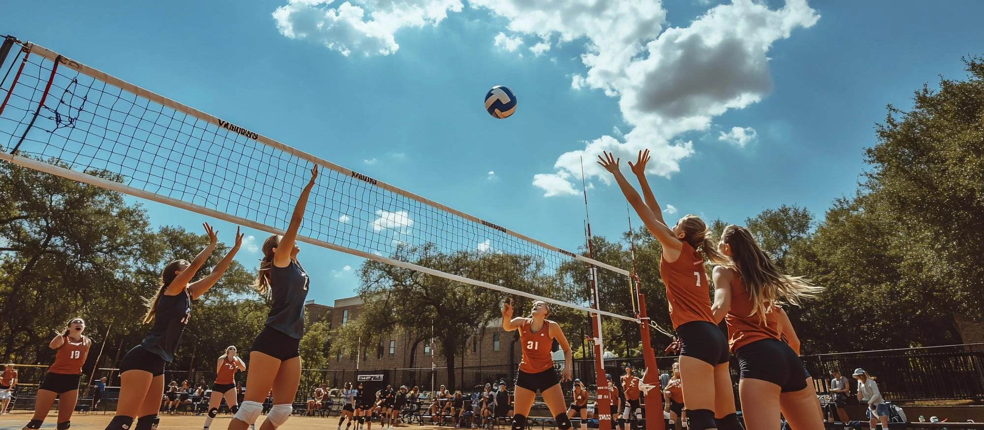 Women playing volleyball outdoors on a sunny day, with some players jumping to hit the ball over the net, surrounded by trees and spectators.