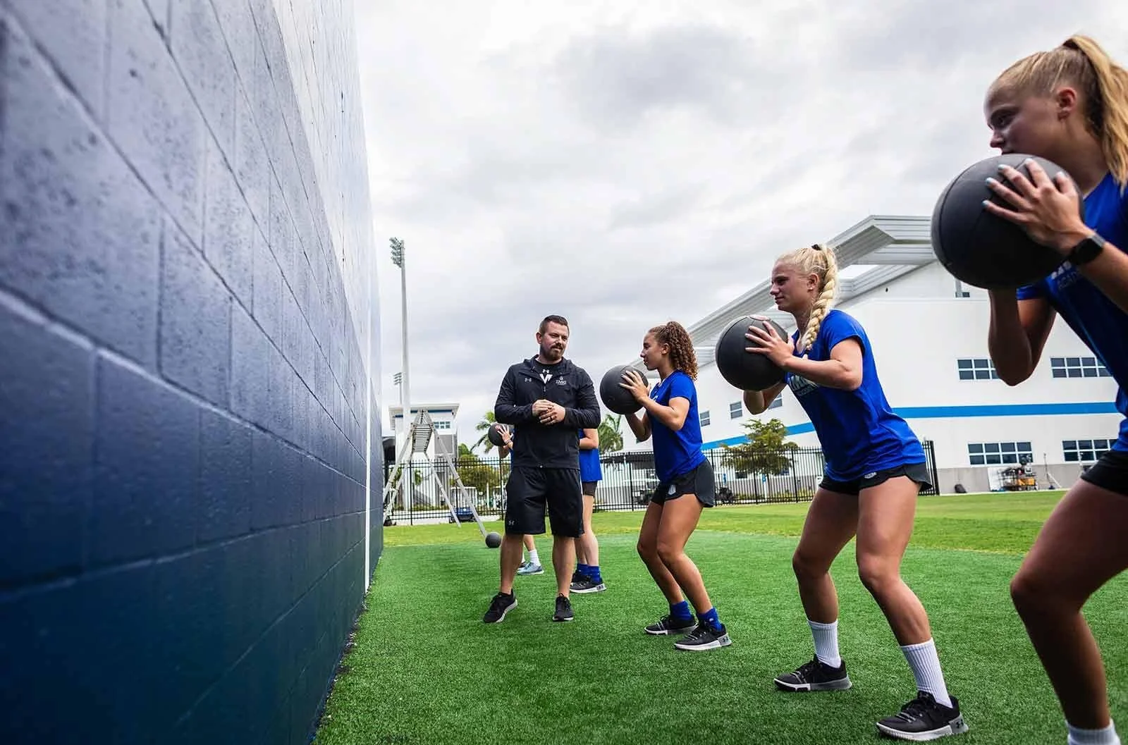 Group of young women in blue sports shirts and black shorts performing wall balls while an instructor supervises, on a grassy outdoor sports field with a large blue wall and a white sports facility in the background.