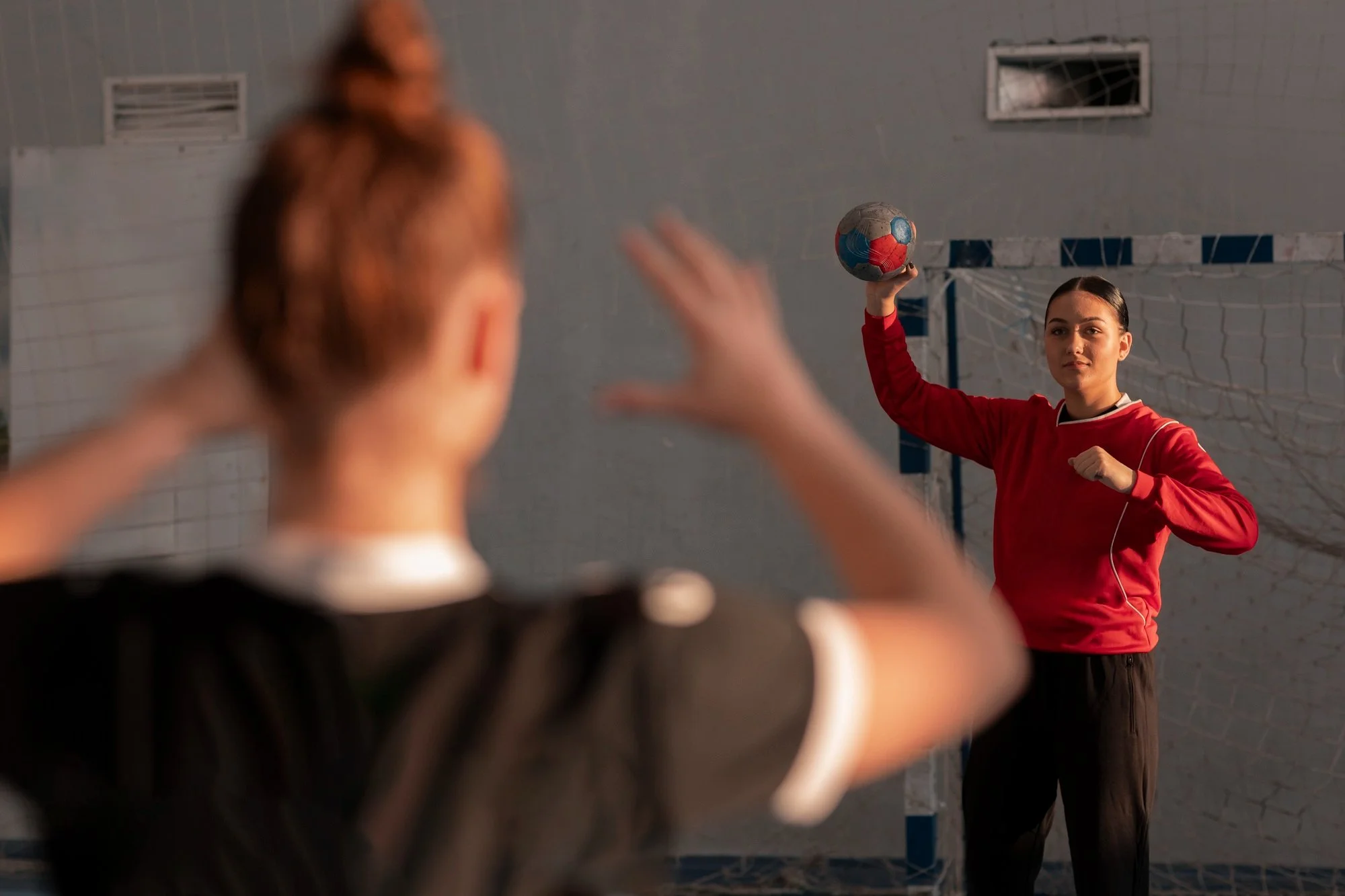 A female soccer player in a red jersey is holding a soccer ball above her head, preparing to throw it in during a game or practice. The photo is taken from behind another player, whose blurred head and hand are visible in the foreground.