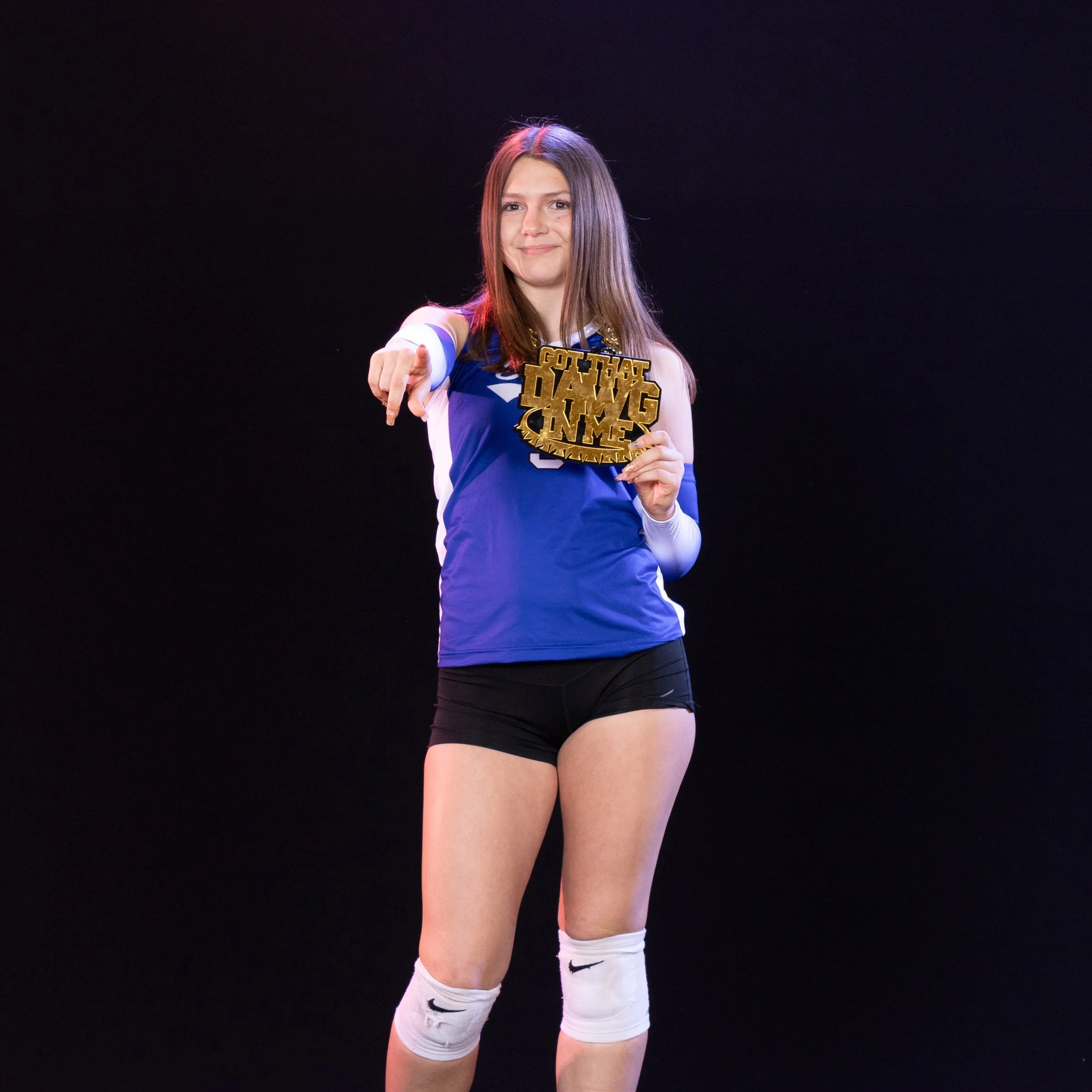 A female athlete in a blue sports uniform and white knee-high socks stands against a black background, smiling and pointing forward while holding a gold medal in her left hand.