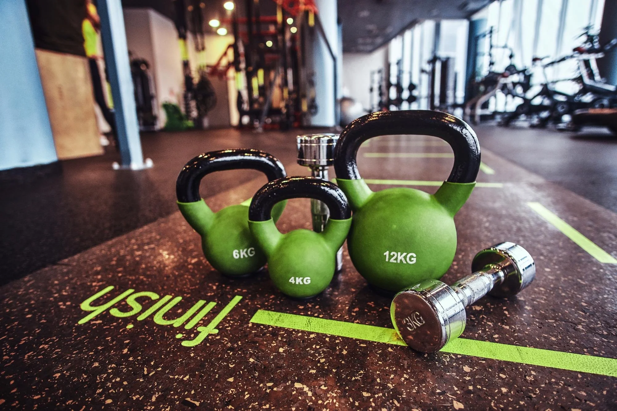 Green kettlebells labeled with weights 4KG, 6KG, and 12KG arranged on a gym floor, with a dumbbell nearby, in a gym setting with workout equipment in the background.