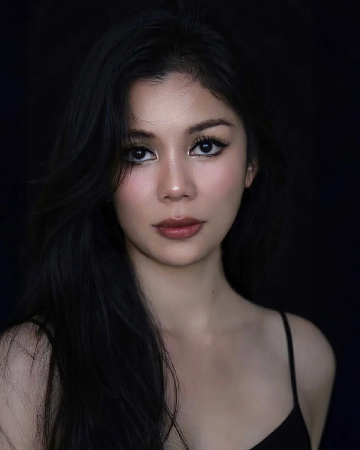 Portrait of a young woman DJ with long dark hair, wearing a black spaghetti strap top, against a black background.