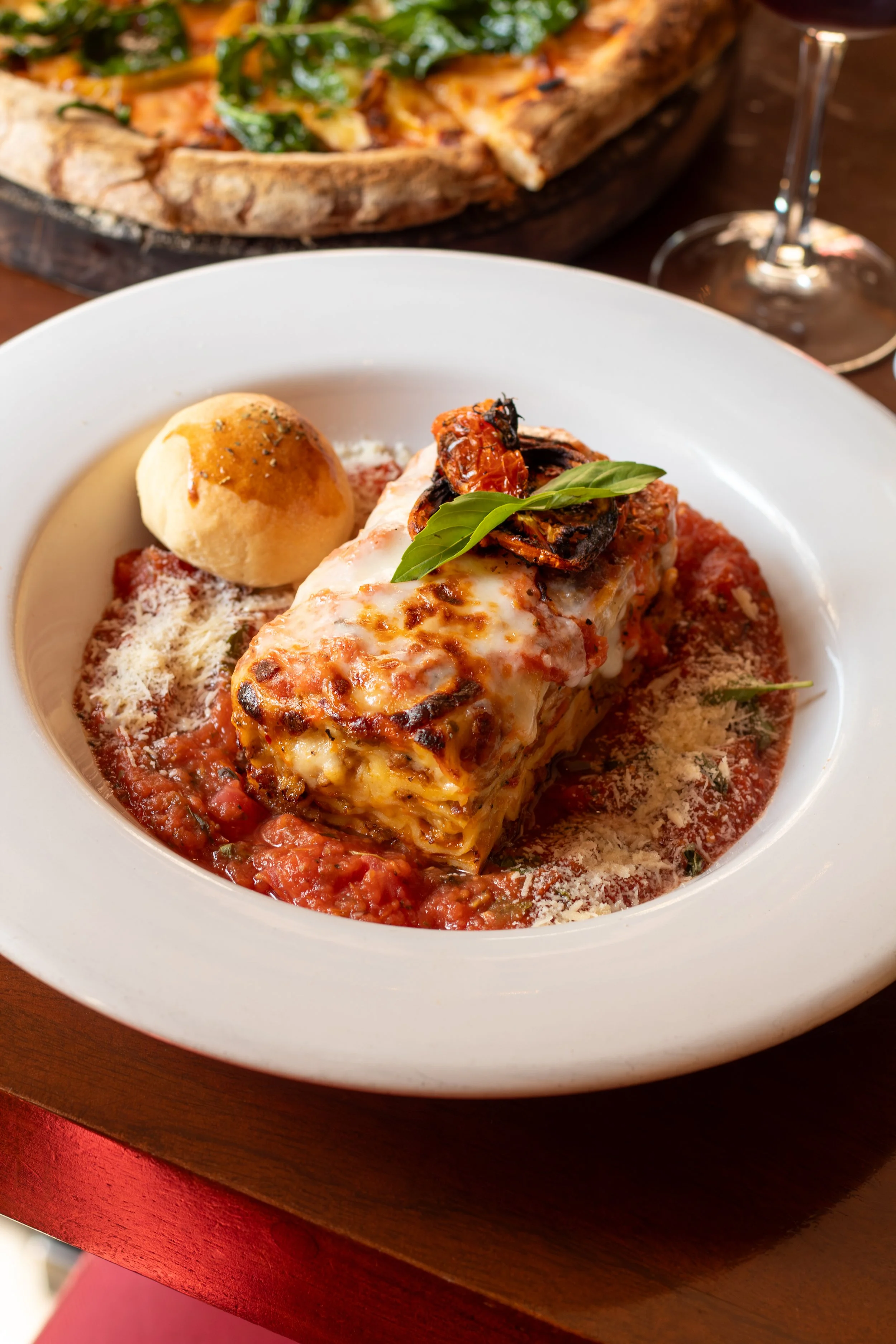Lasagna de carne con salsa de tomate, queso derretido, albahaca y una bola de pan. En el fondo se ve una pizza al horno de leña en una bandeja de madera.