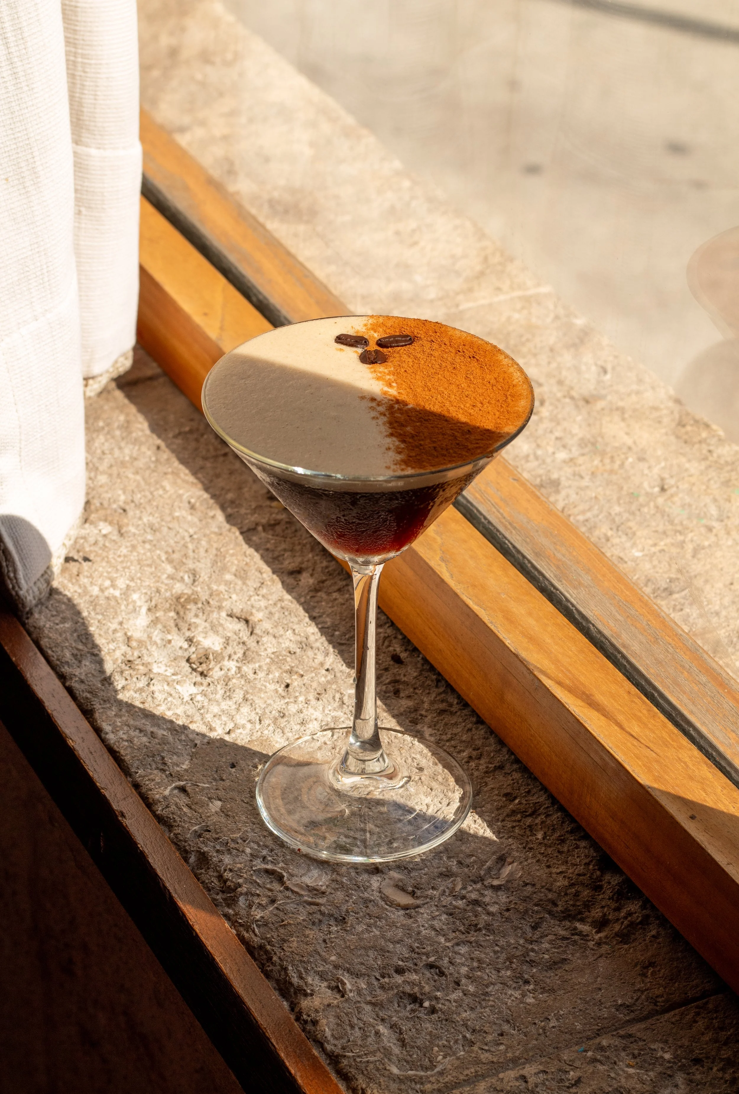 Espresso Martini. Cóctel oscuro en copa de cristal sobre mesón de piedra, decorado con polvo de cacao y granos de café.