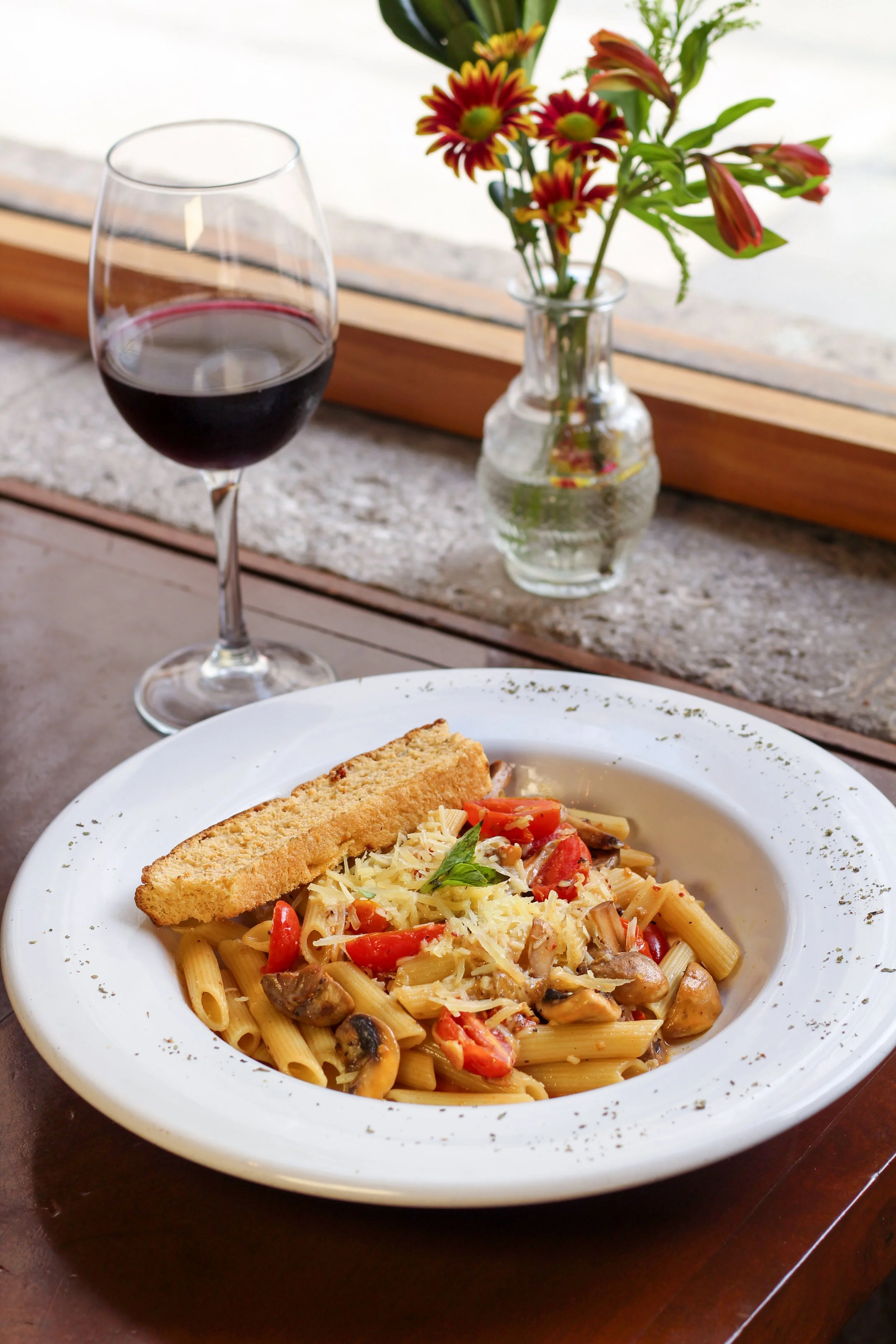 Penne al Aglio y Peperoncino con Hongos Rostizados. Plato de pasta con vegetales, cubierto con queso rallado y pan frito, junto a una copa de vino tinto, en un restaurante con flores en una jarra y vista al exterior.