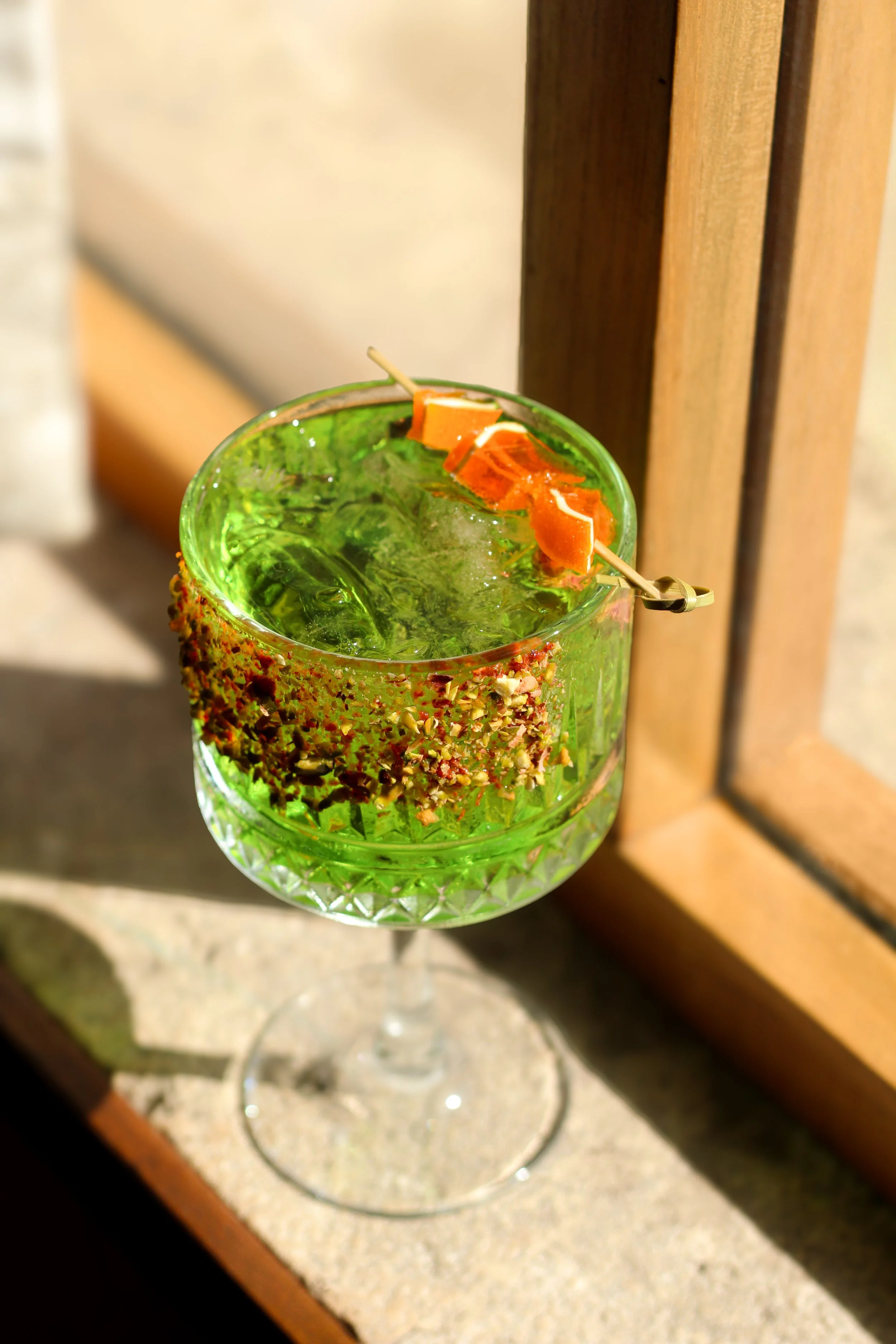 Cóctel verde en copa de cristal con borde decorado con semillas, servido con hielo y una rodaja de naranja, en un ambiente iluminado y decorado.