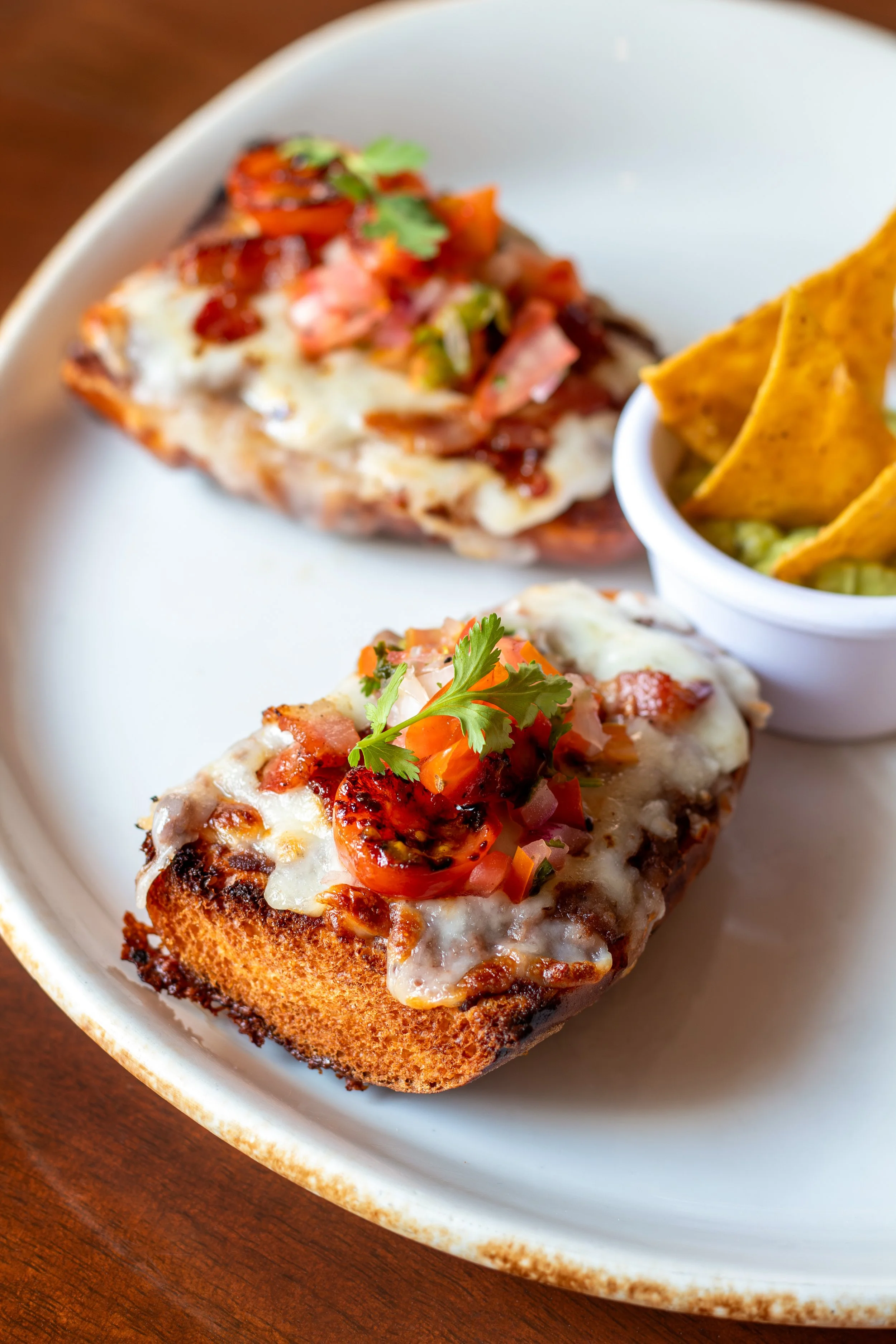 Molletes con tocino, servidos con queso, tomate y cilantro en plato blanco sobre mesa de madera.