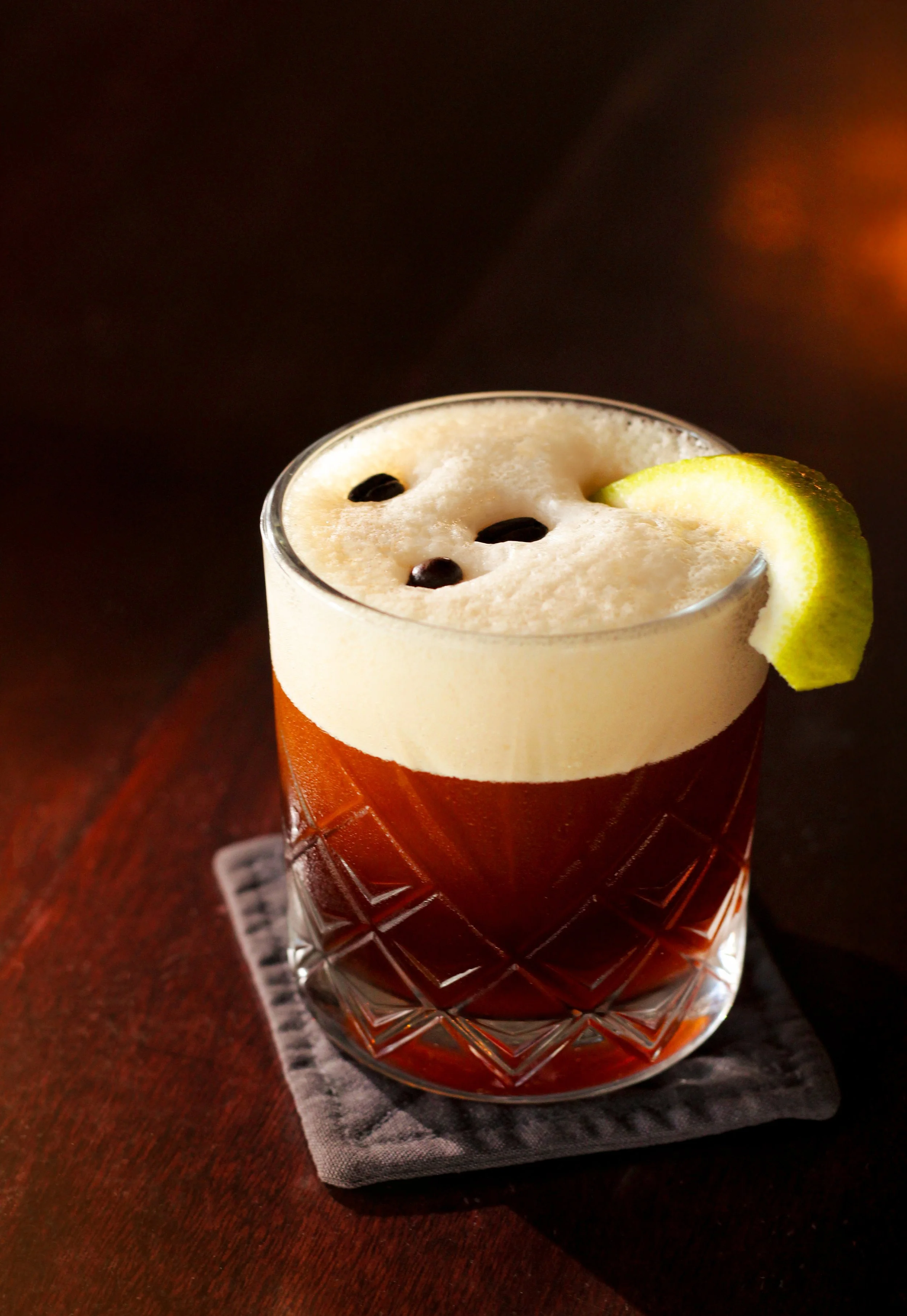 Carajillo shakeado. Coctel a base de café y Licor 43 en vaso decorado con una rodaja de limón y hielo, en una mesa de madera con fondo oscuro.