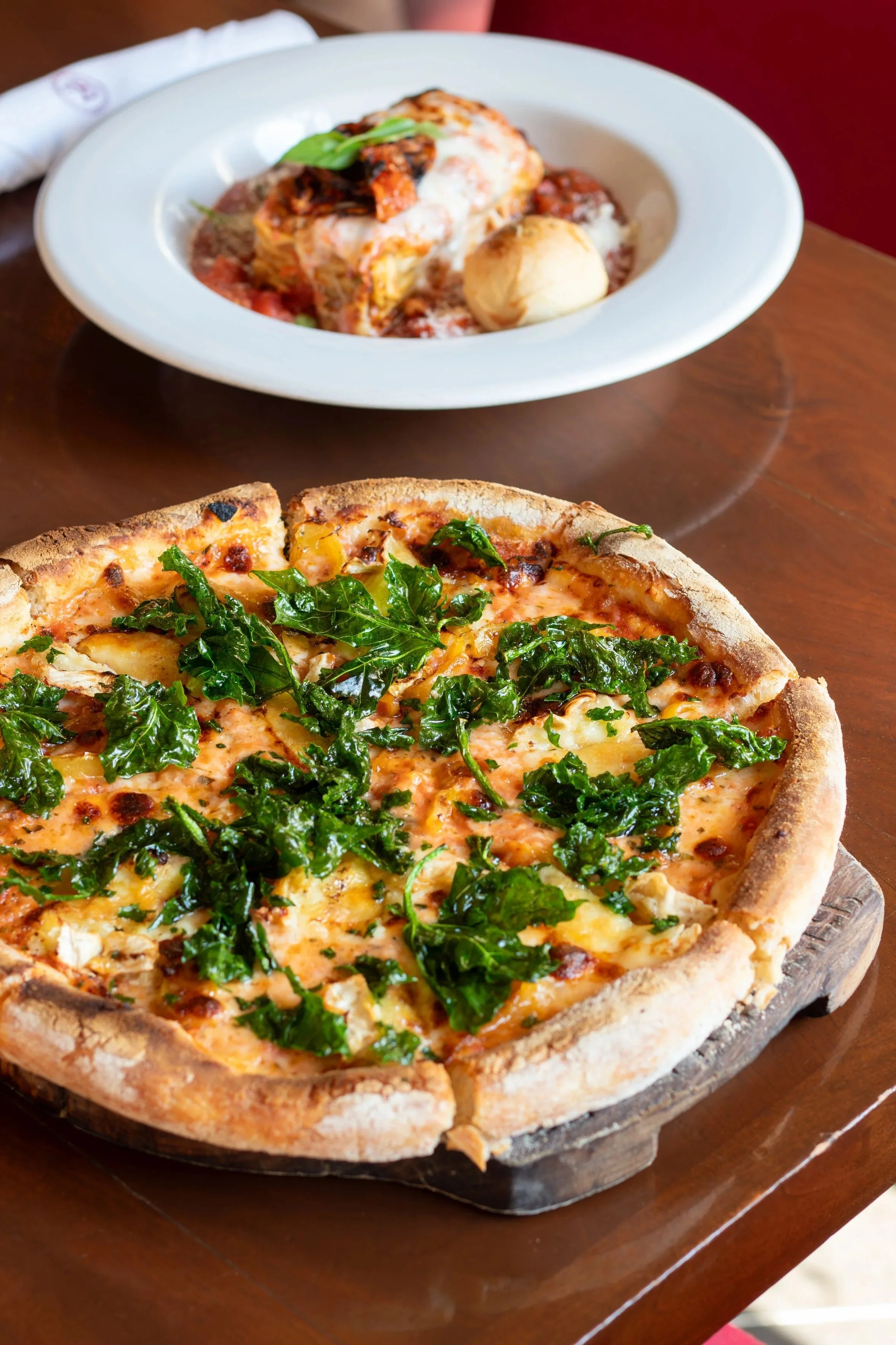 Pizza al horno de leña de Chaya Frita, Chile Xcatic y Queso Brie. Una pizza con queso, espinacas y otras verduras, sobre una tabla de madera, en primer plano.