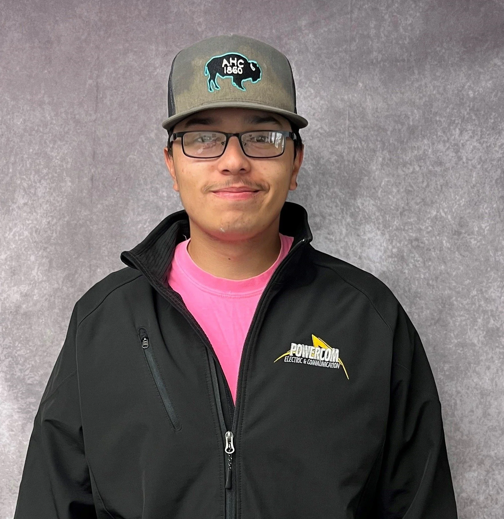 A young man wearing glasses, a gray and black snapback hat with a neon outline of a buffalo and the text 'AHC 1860', a pink shirt, and a black jacket with a yellow and white 'Powercom Electric & Communication' logo, standing against a gray wall.