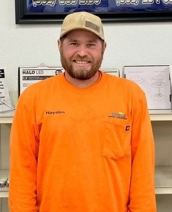 A man with a beard wearing an orange long-sleeve shirt and beige cap, standing indoors in front of shelves with boxed products.