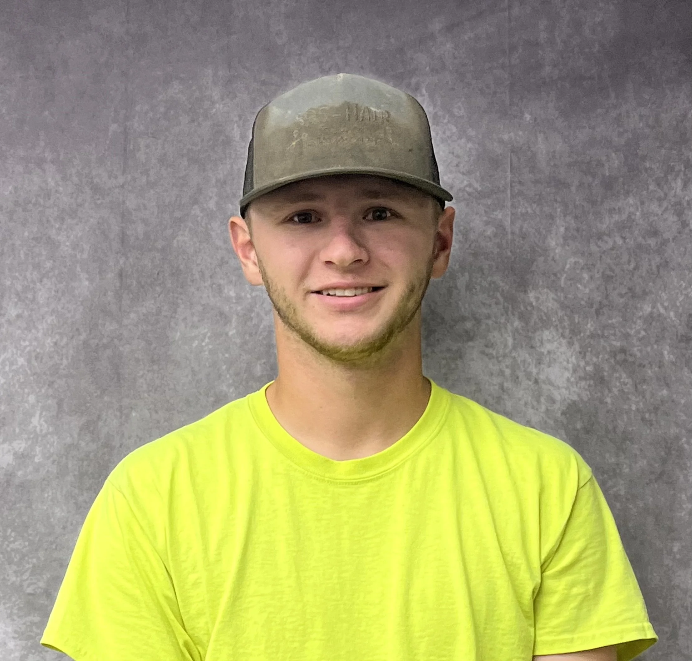 A young man smiling, wearing a neon yellow shirt and a camo baseball cap, standing against a gray background.