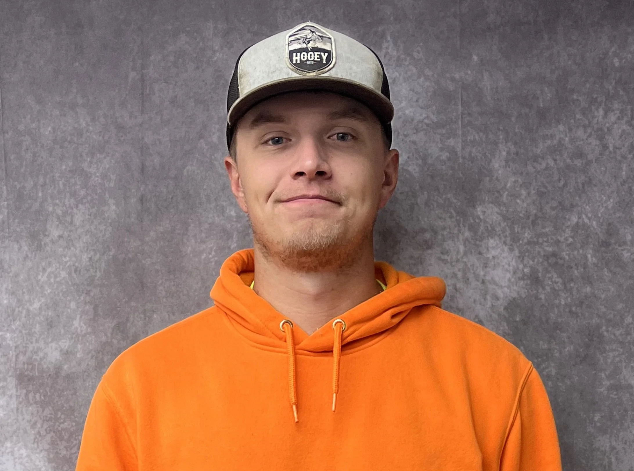 A young man with light skin, blue eyes, and a short beard is wearing an orange hoodie and a gray baseball cap with a black front panel, standing against a gray wall.