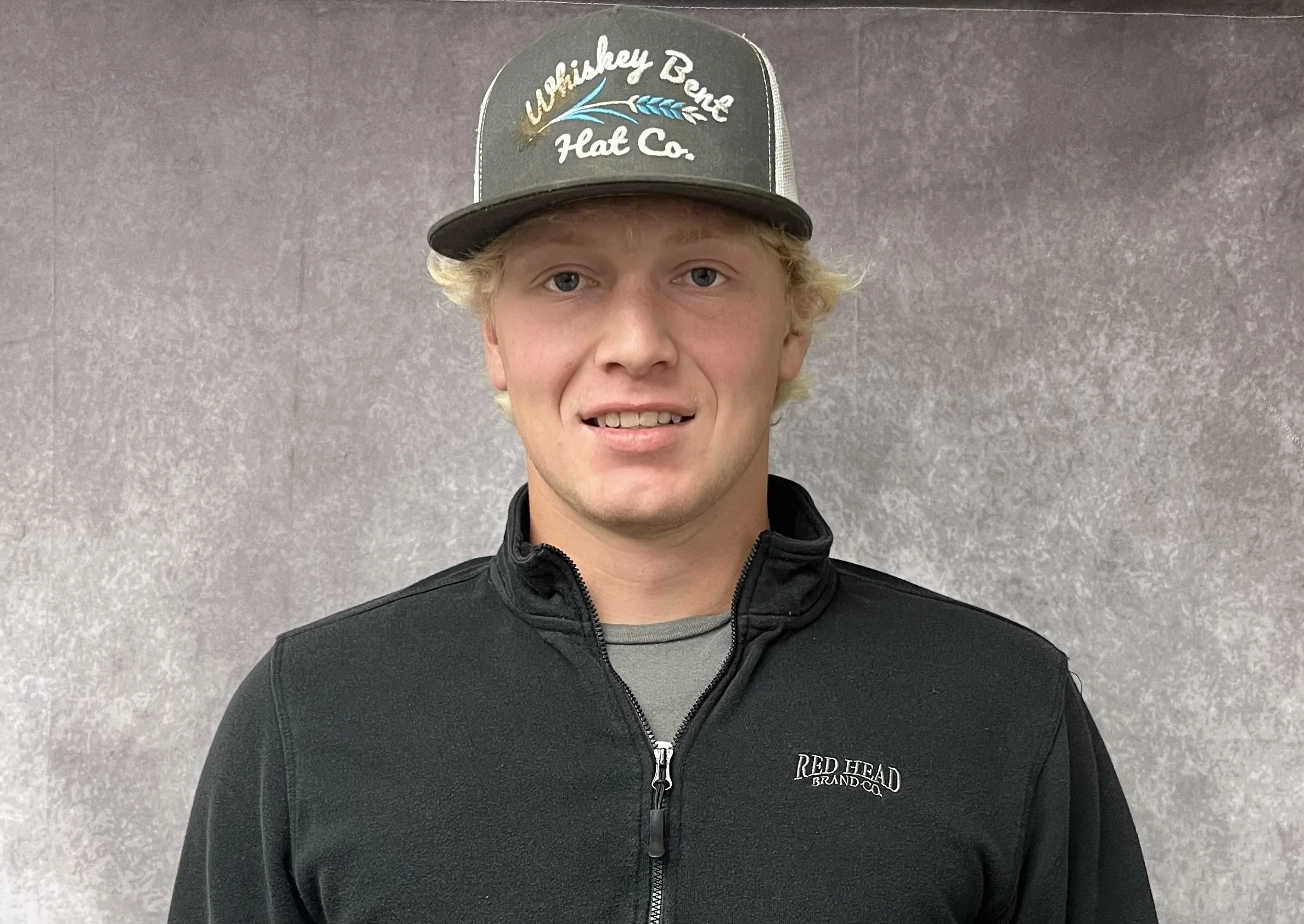 A young man with blonde hair wearing a gray and white trucker hat with the logo 'Whiskey Bend Flat Co.', a gray shirt, and a black zip-up jacket with the words 'Red Head Brand Co.' on it, standing in front of a gray textured wall.