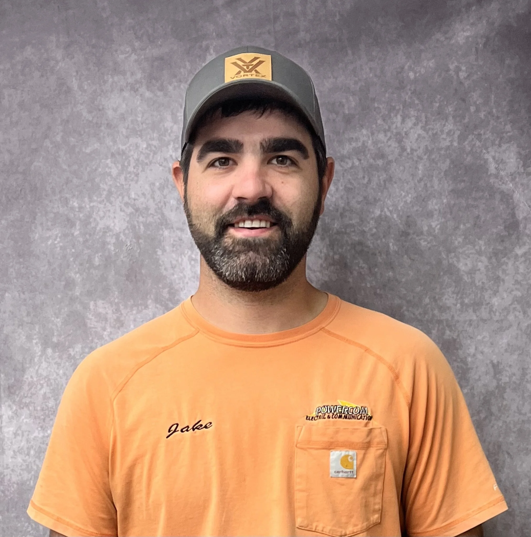 A man with dark hair and beard wearing an orange Carhartt T-shirt and a gray cap, standing against a gray textured background.