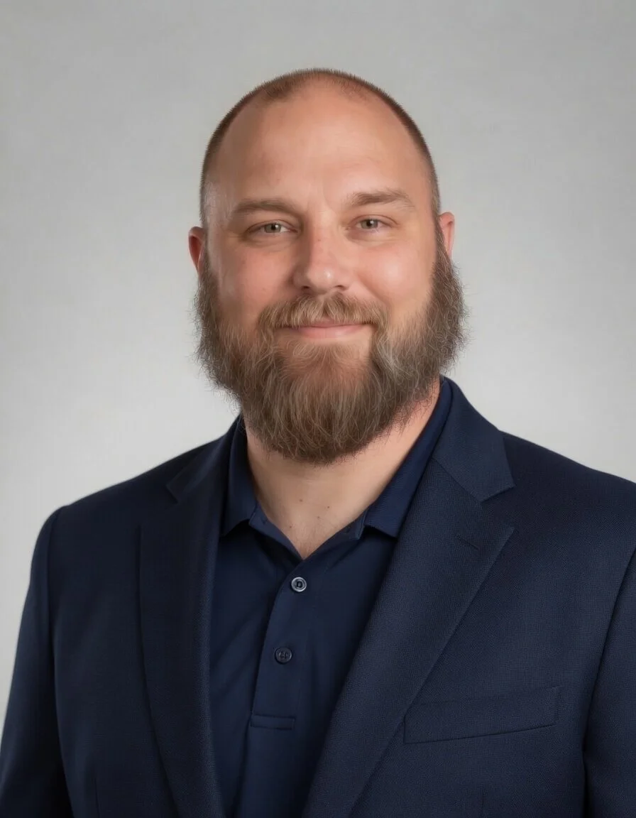 A man with a beard and short hair wearing a dark blue suit and shirt, smiling at the camera against a plain light background.
