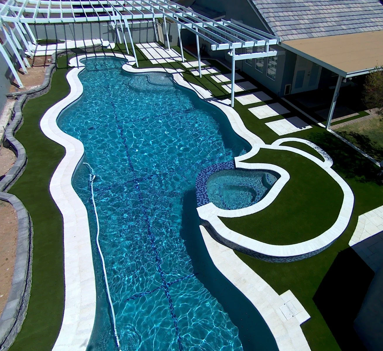 A backyard swimming pool with a unique freeform shape, surrounded by a white concrete deck and green artificial grass. There is a small circular hot tub attached to the pool. The pool has a blue mosaic tile border on one side. Nearby is a covered patio area with a roof supported by white columns, and there are lounge chairs and potted plants around.