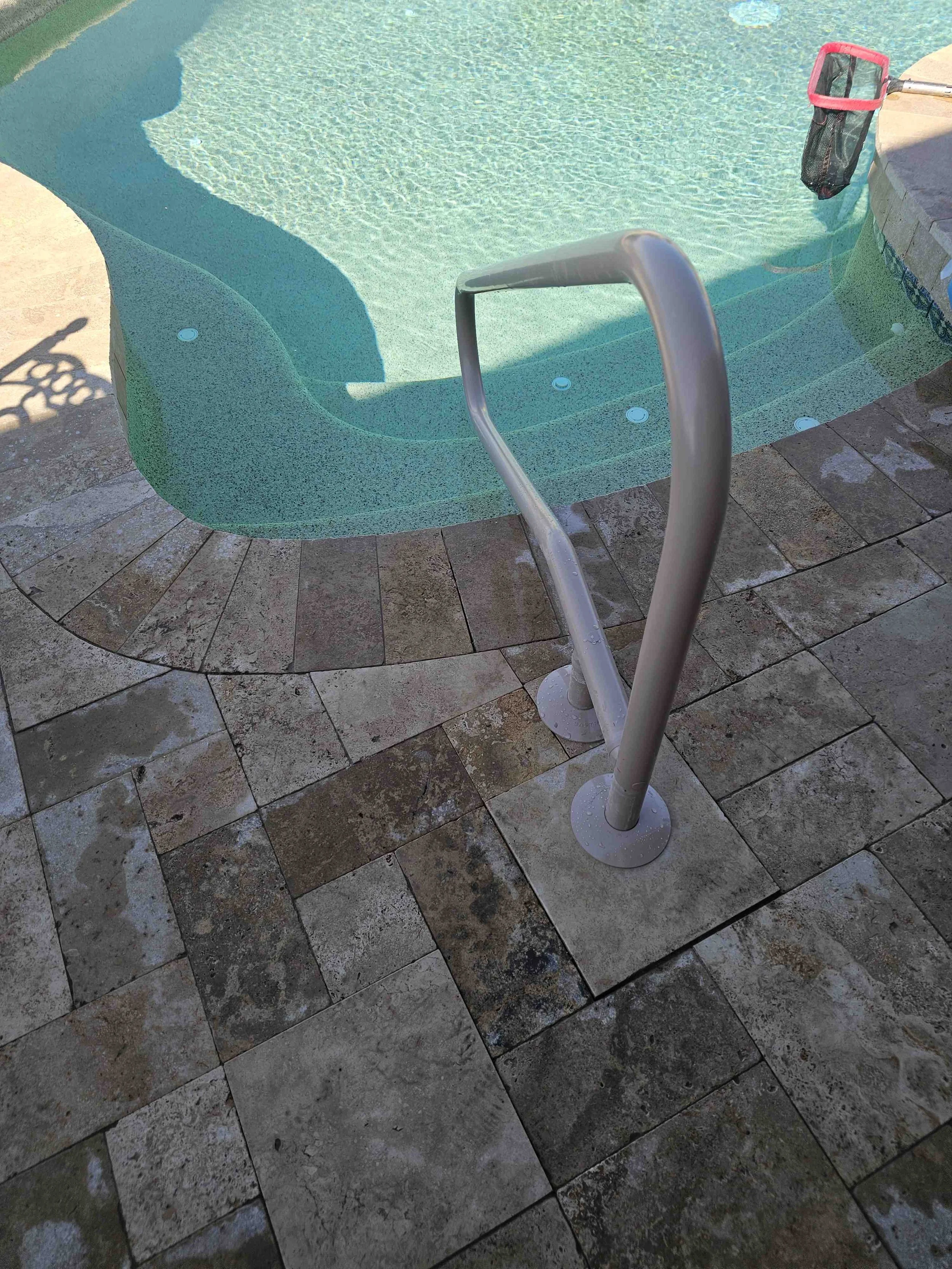 Backyard swimming pool with steps, metal pool ladder, a pool skimmer net hanging on the side, and a stone deck surrounding the pool.