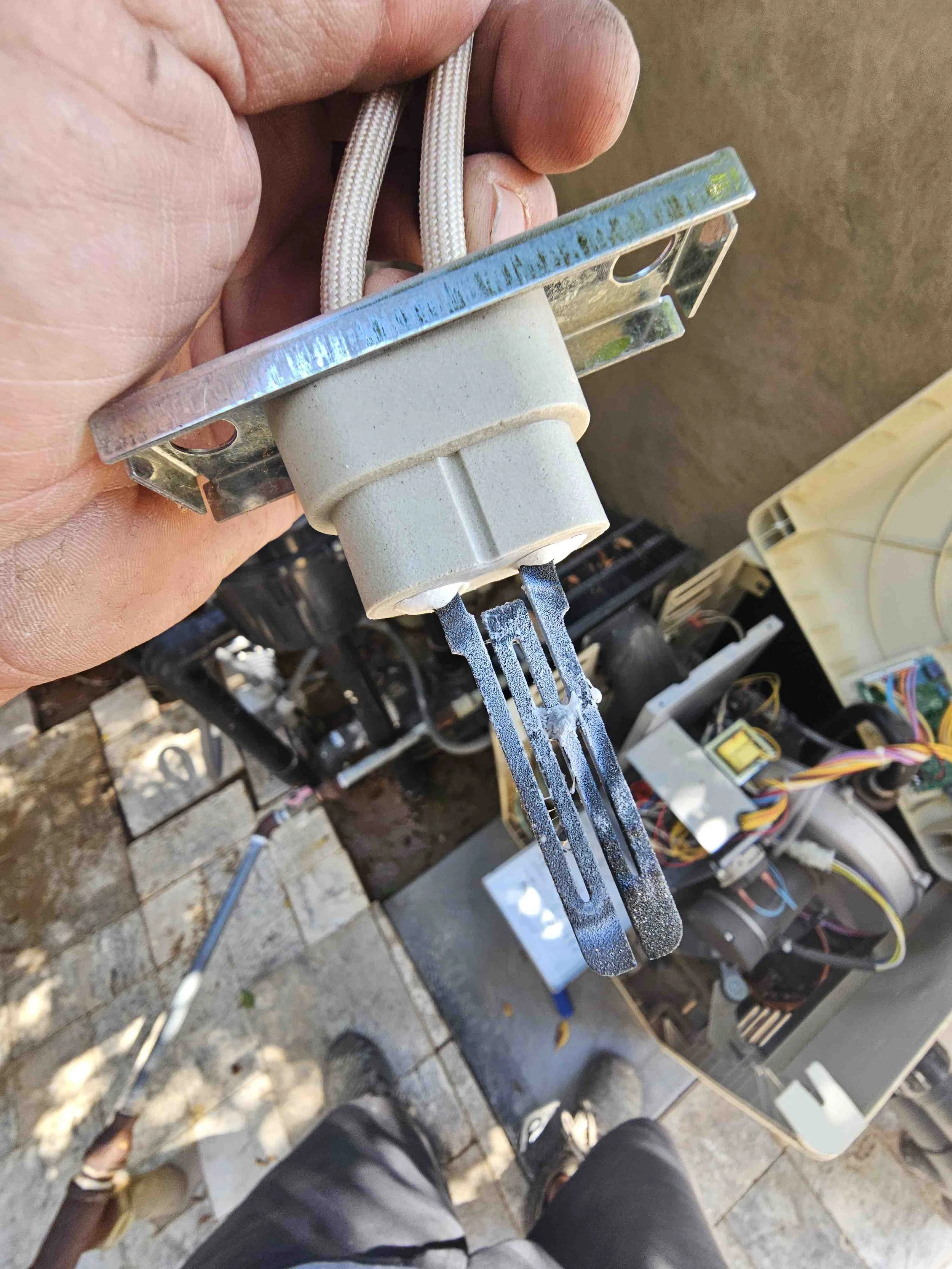 Close-up of a person's hand holding a metal electrical or electronic component with wires and circuit boards in the background.