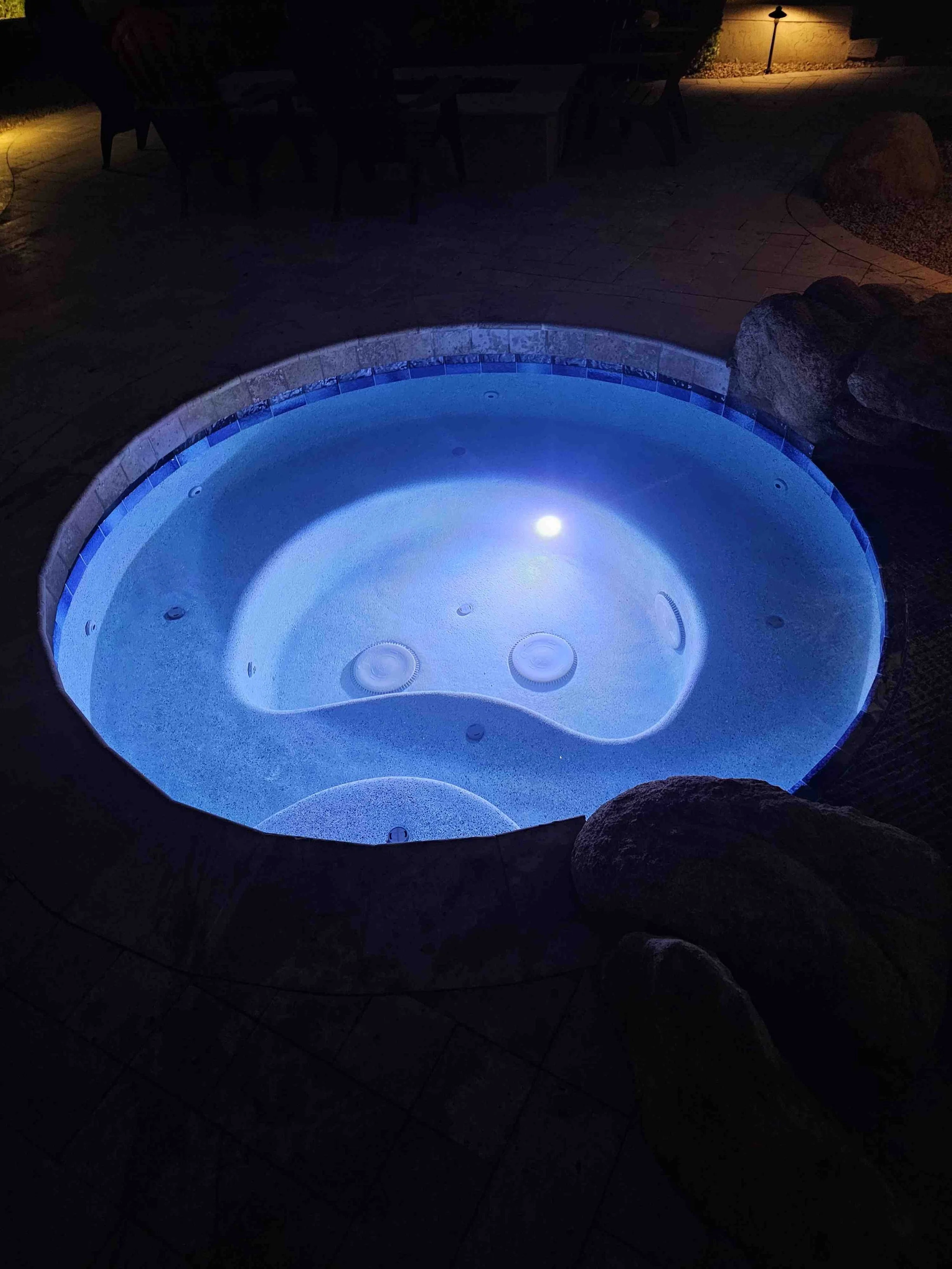 A hot tub illuminated with blue LED lights in an outdoor patio at night, surrounded by rocks and patio furniture.