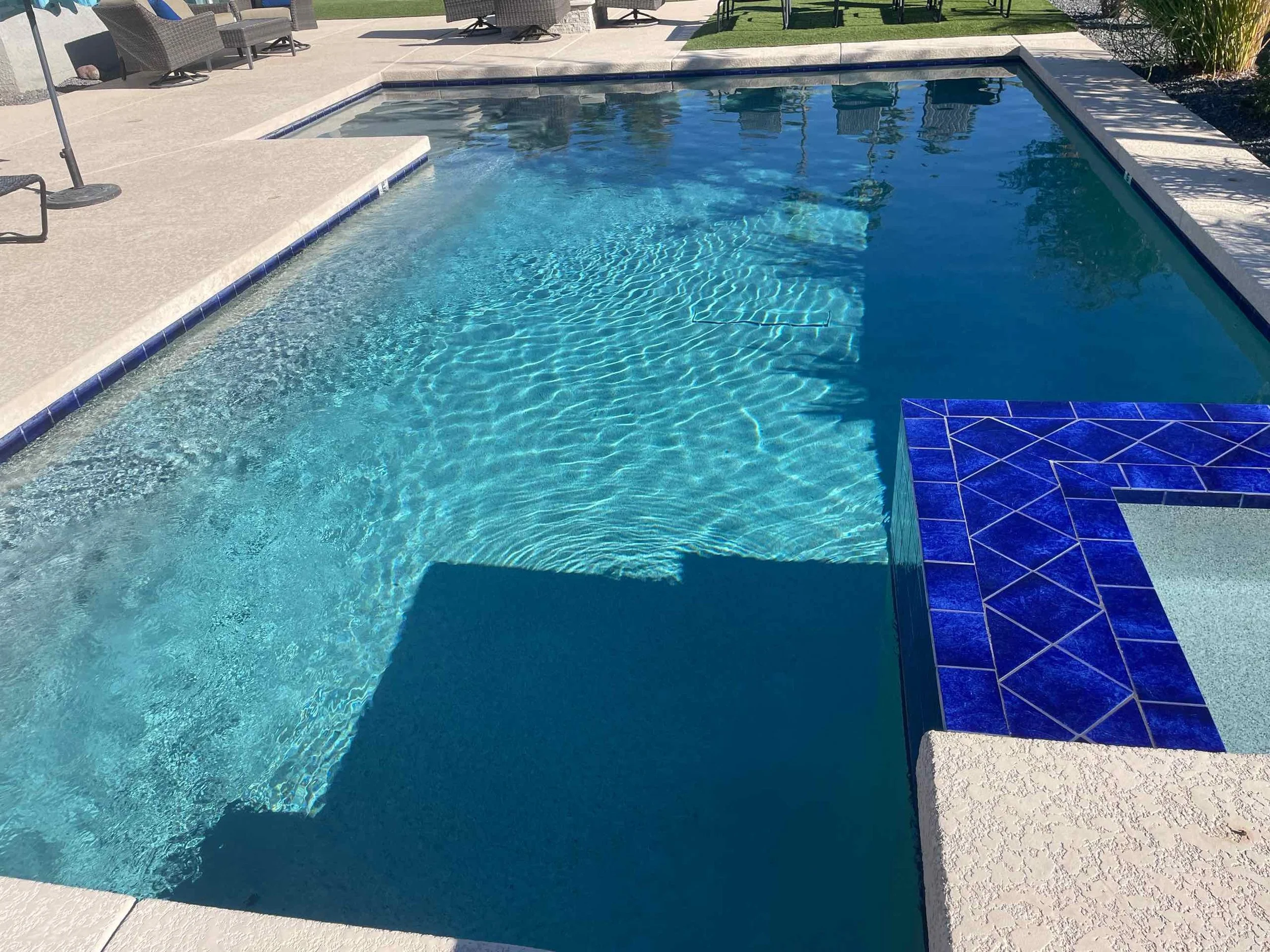 A backyard swimming pool with clear blue water, surrounded by concrete and tiled edges, with patio furniture and plants in the background.